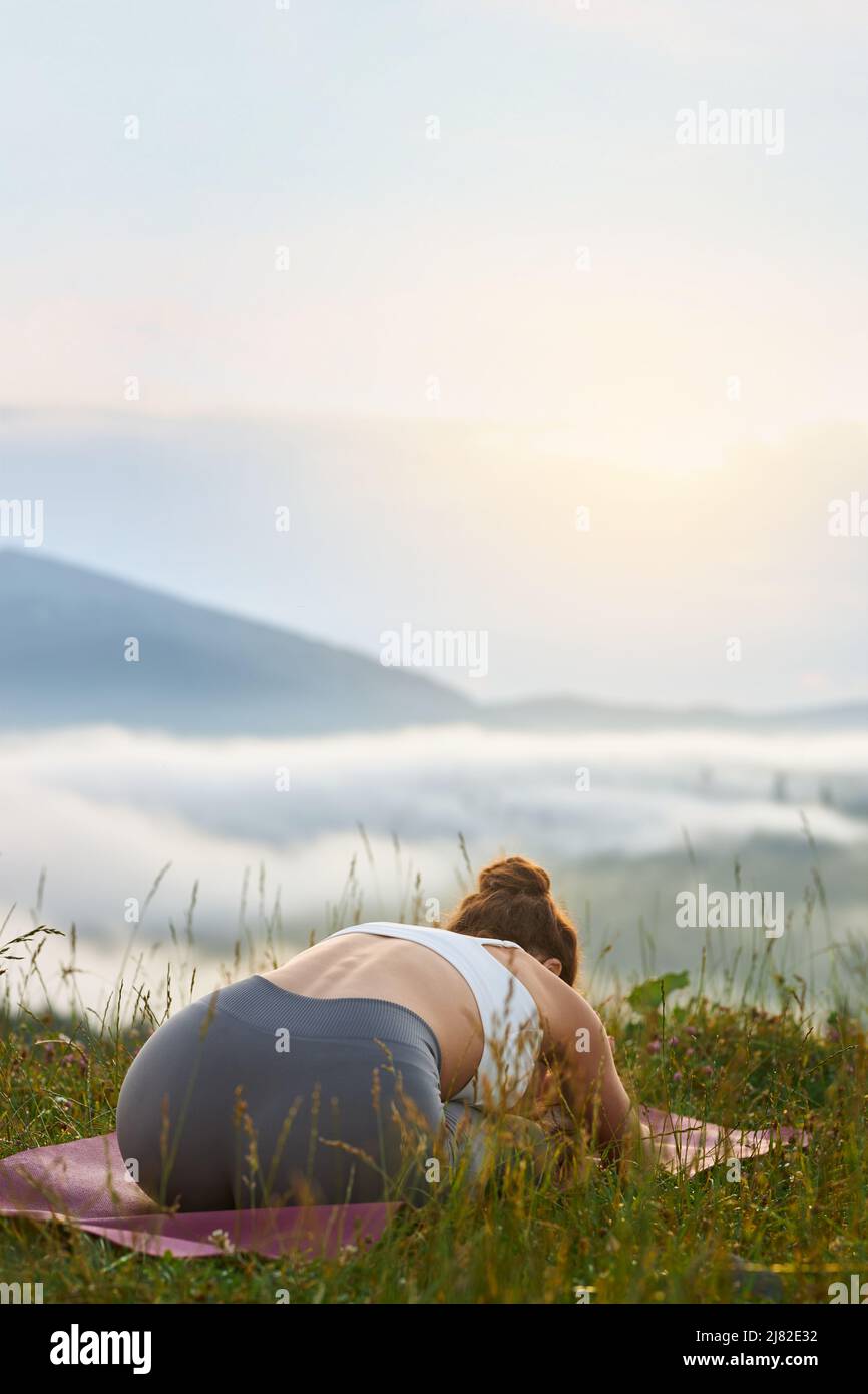 Rückansicht einer jungen schlanken Frau, die in Hügeln Yoga praktiziert. Flexible weibliche sitzende auf Beinen, die sich in den Bergen auf einer Yogamatte beugte. Konzept der Religion des neuen Zeitalters. Stockfoto