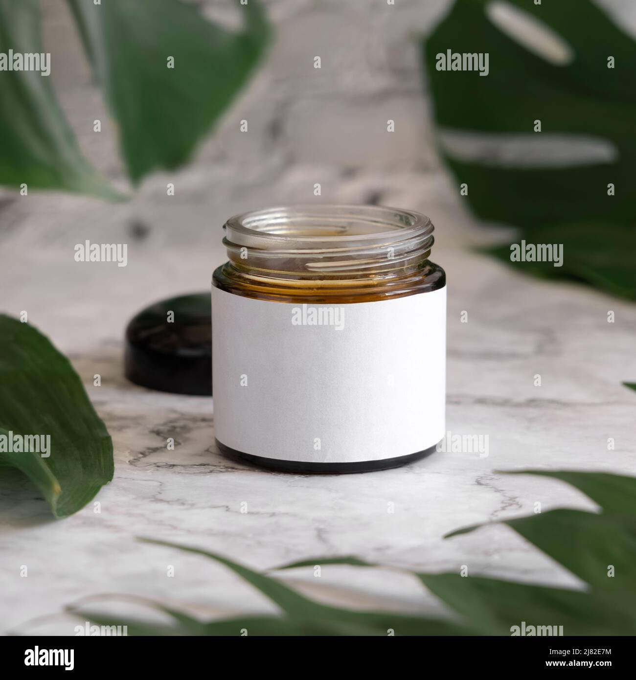 Geöffnetes braunes Glas und Monstera-Blätter auf Marmortisch aus nächster Nähe. Modell der Markenverpackung. Hautpflegeprodukt, Creme oder Butter. Hausgemachte Herba Stockfoto