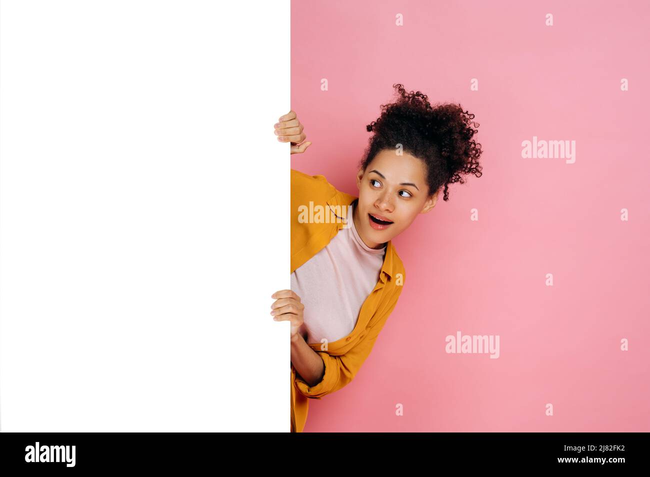 Staunendes afroamerikanisches Mädchen mit lockigem Haar, das aus dem weißen Brett mit Mockup-Schablone und Kopieplatz für Präsentation oder Werbung guckt, auf rosa Hintergrund steht und lächelt. Modellkonzept Stockfoto