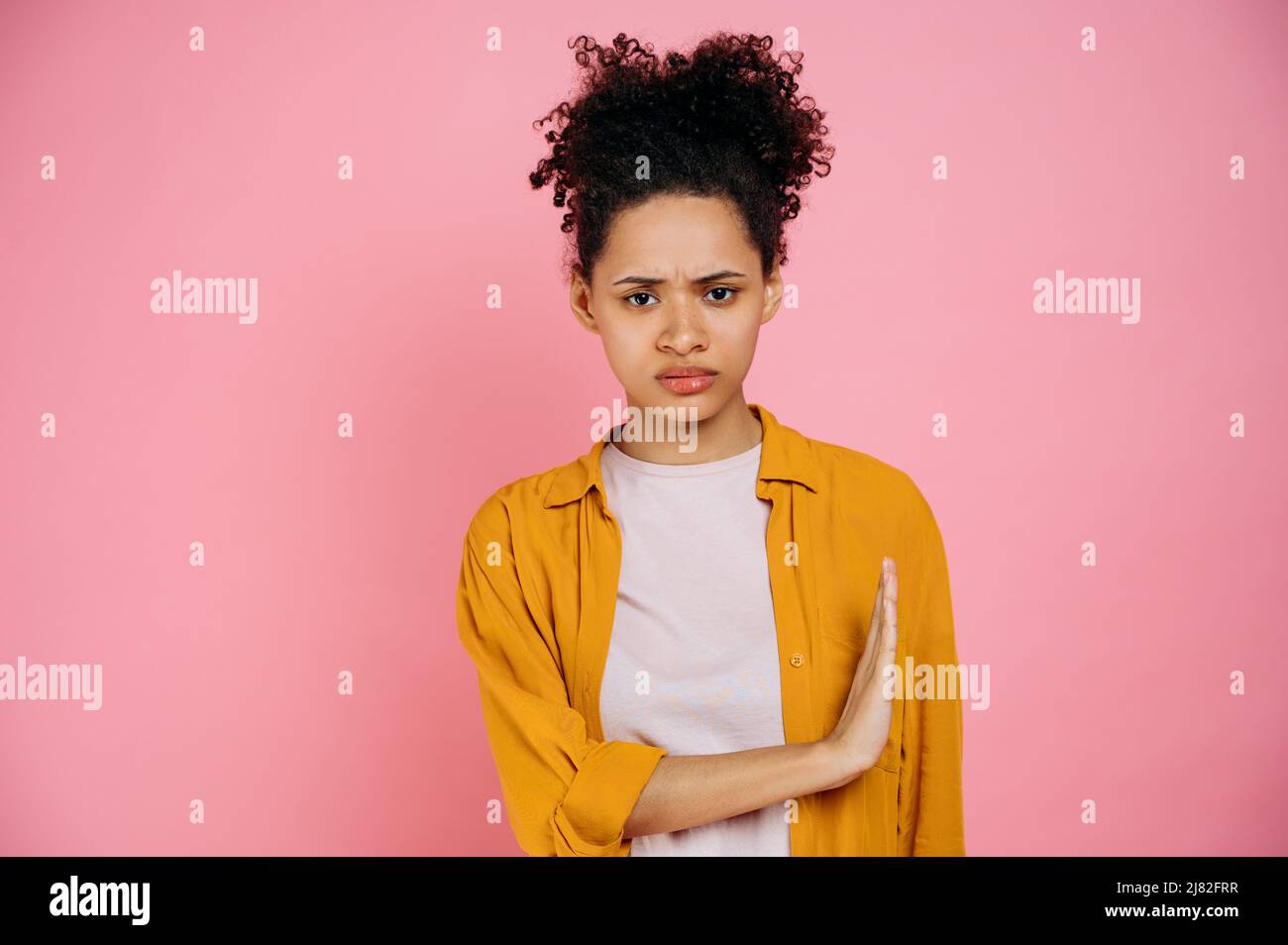 Trauriges, lockiges afroamerikanisches Mädchen in legerer Kleidung, das Nein sagt, zeigt eine Stop-Geste mit der Handfläche zur Seite, drückt Unzufriedenheit aus, steht auf einem isolierten rosa Hintergrund und blickt auf die Kamera Stockfoto