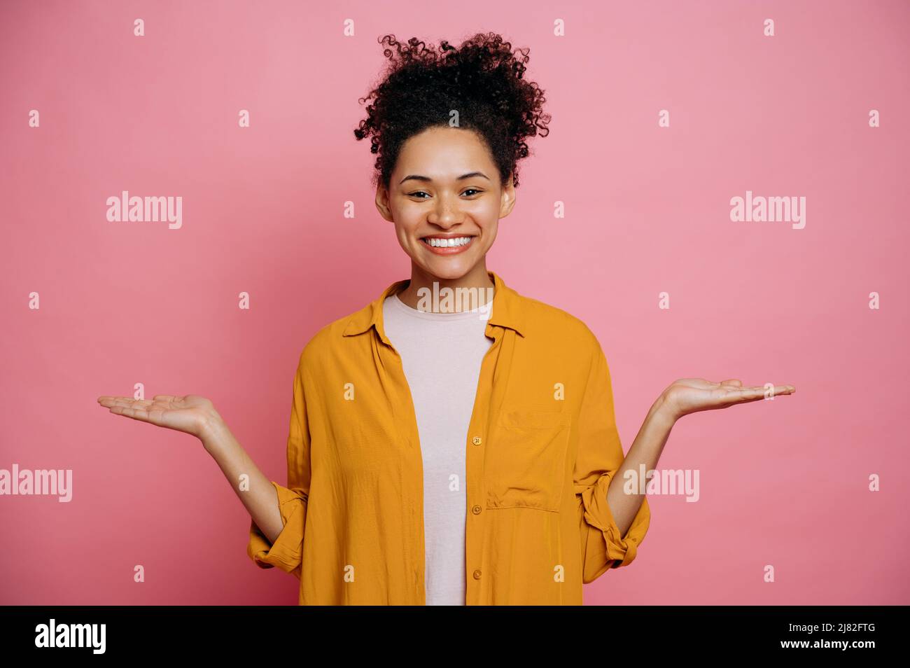 Fröhliches afroamerikanisches Mädchen mit lockigem Haar in legerer Kleidung, das mit den Händen zu den Seiten auf einen leeren Raum für eine Präsentation zeigt, blickt lächelnd auf die Kamera und steht auf einem isolierten rosa Hintergrund Stockfoto