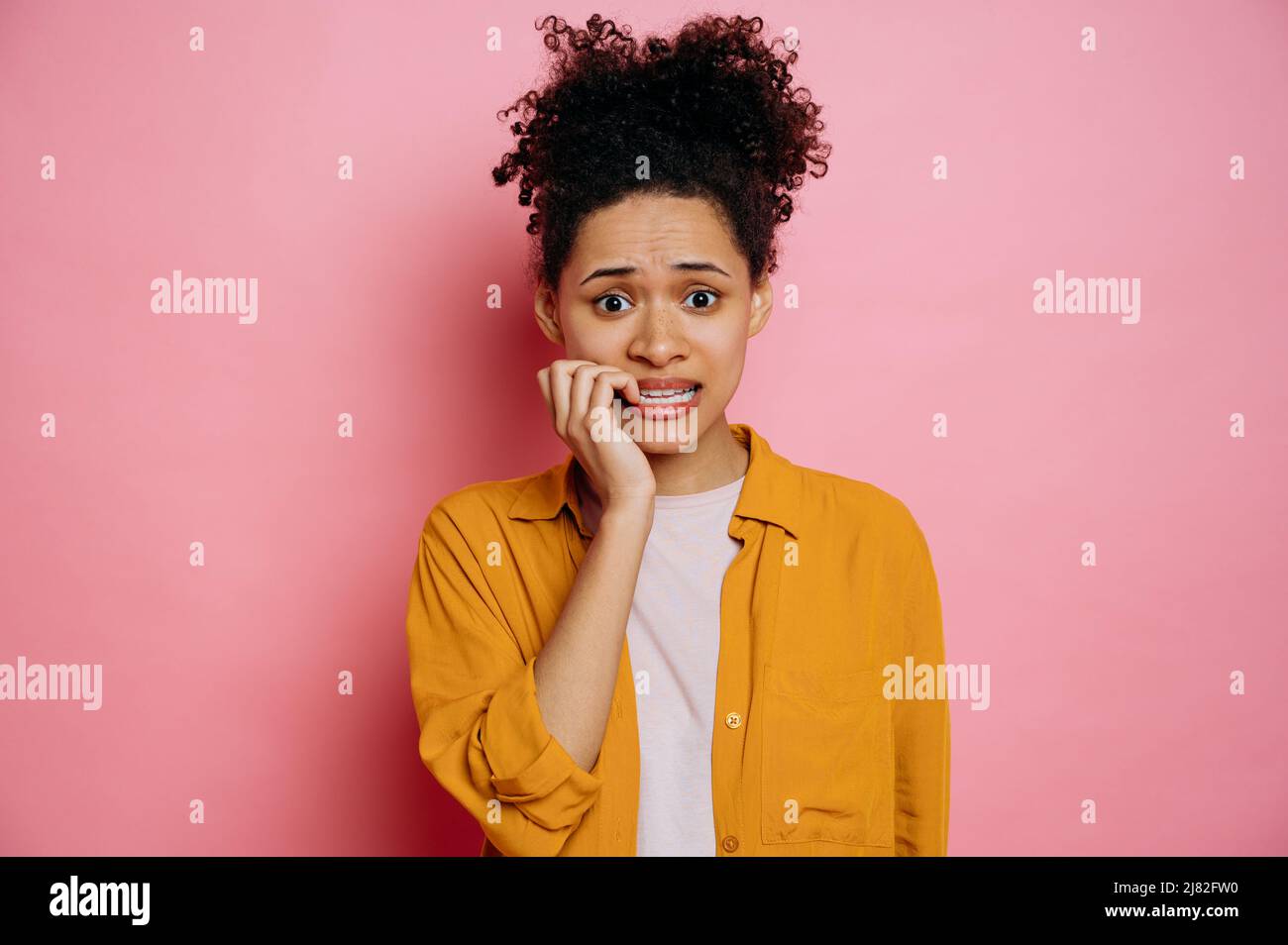 Betäubt verwirrt afroamerikanische junge Frau, in Freizeitkleidung, gestresst und nervös mit Händen auf dem Mund beißende Nägel, Blick auf die Kamera, geht durch, steht auf isoliertem rosa Hintergrund Stockfoto