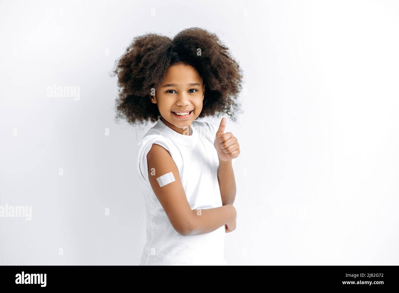 Positives afroamerikanisches kleines Mädchen, Vorschulkind, mit Pfand auf der Schulter nach der Impfung, für Immunität gegen covid19 und andere Krankheiten, stehen auf isoliertem weißen Hintergrund, Daumen-up-Geste Stockfoto