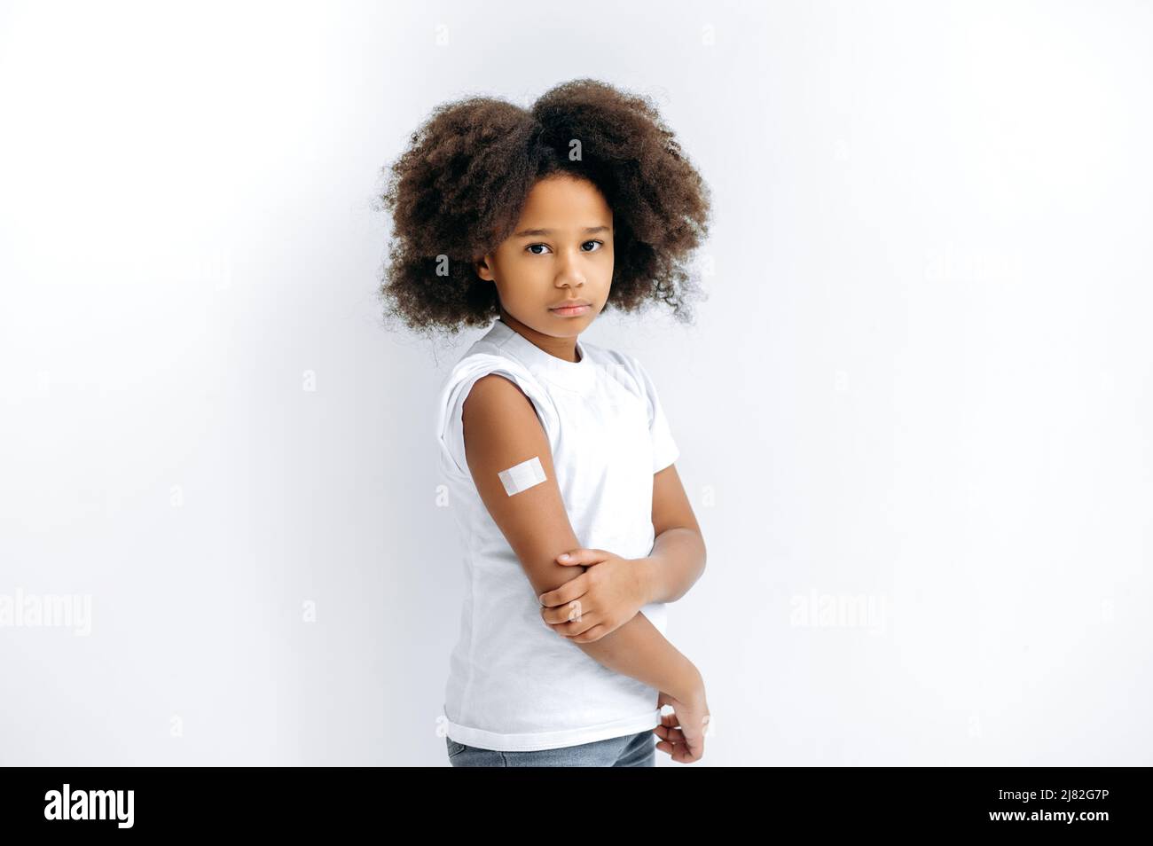 Verärgert afroamerikanisch lockiges Mädchen, das nach der Impfung ein Pfand auf der Schulter hat, steht auf einem isolierten weißen Hintergrund und schaut in die Kamera. Virenschutz, Immunitätskonzept Stockfoto