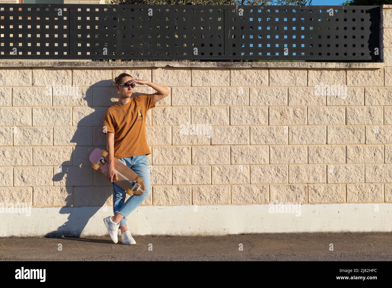 Skater Mann entspannt hält sein Longboard an einer weißen Wand direkt auf das Sonnenlicht vor Sonnenuntergang Zeit schauen Schatten auf Gesicht Stockfoto