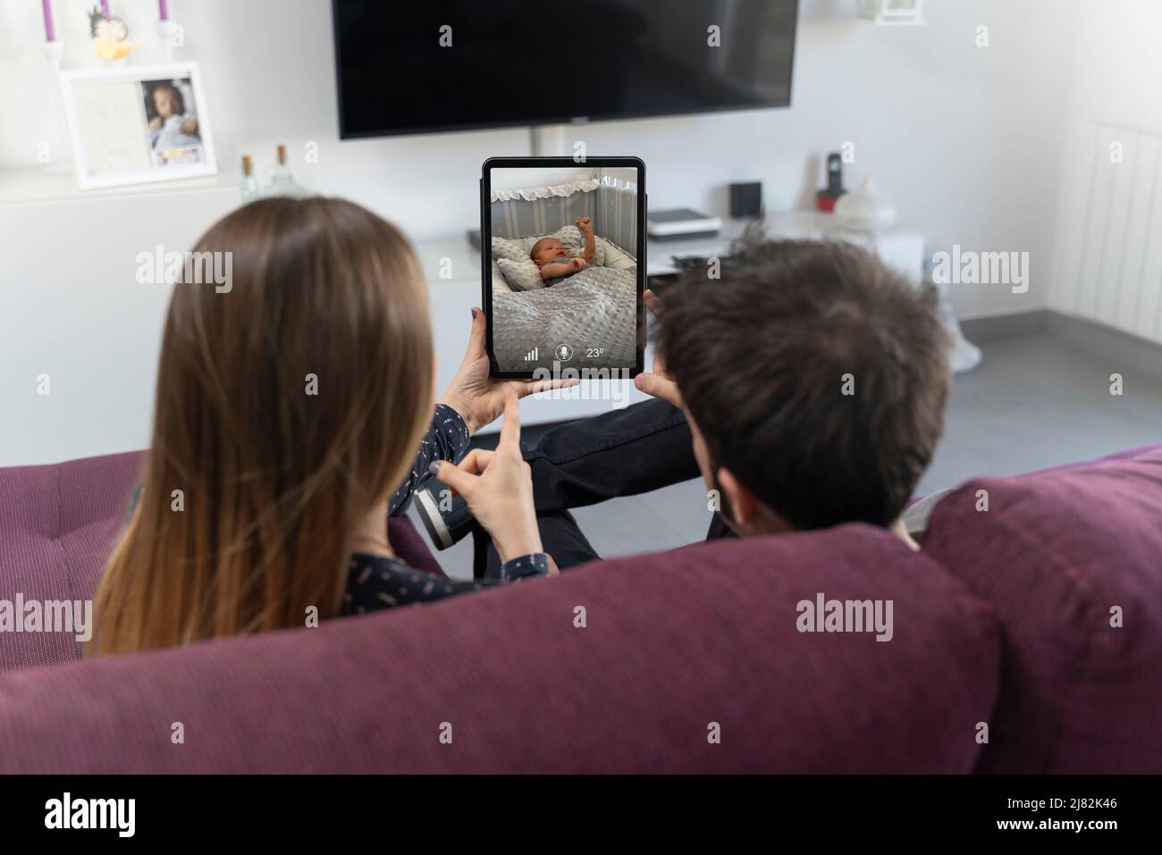 Zwei Eltern sitzen auf dem Sofa und überwachen ihr Baby auf dem Tablet Stockfoto