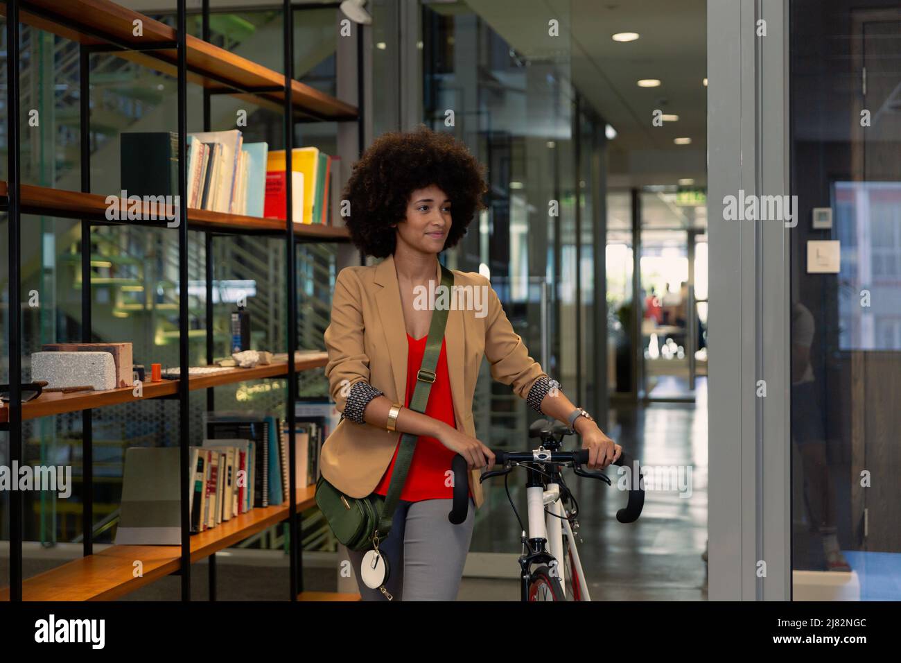 Lächelnde afroamerikanische junge Geschäftsfrau mit Afro-Frisur watend Fahrrad am modernen Arbeitsplatz Stockfoto