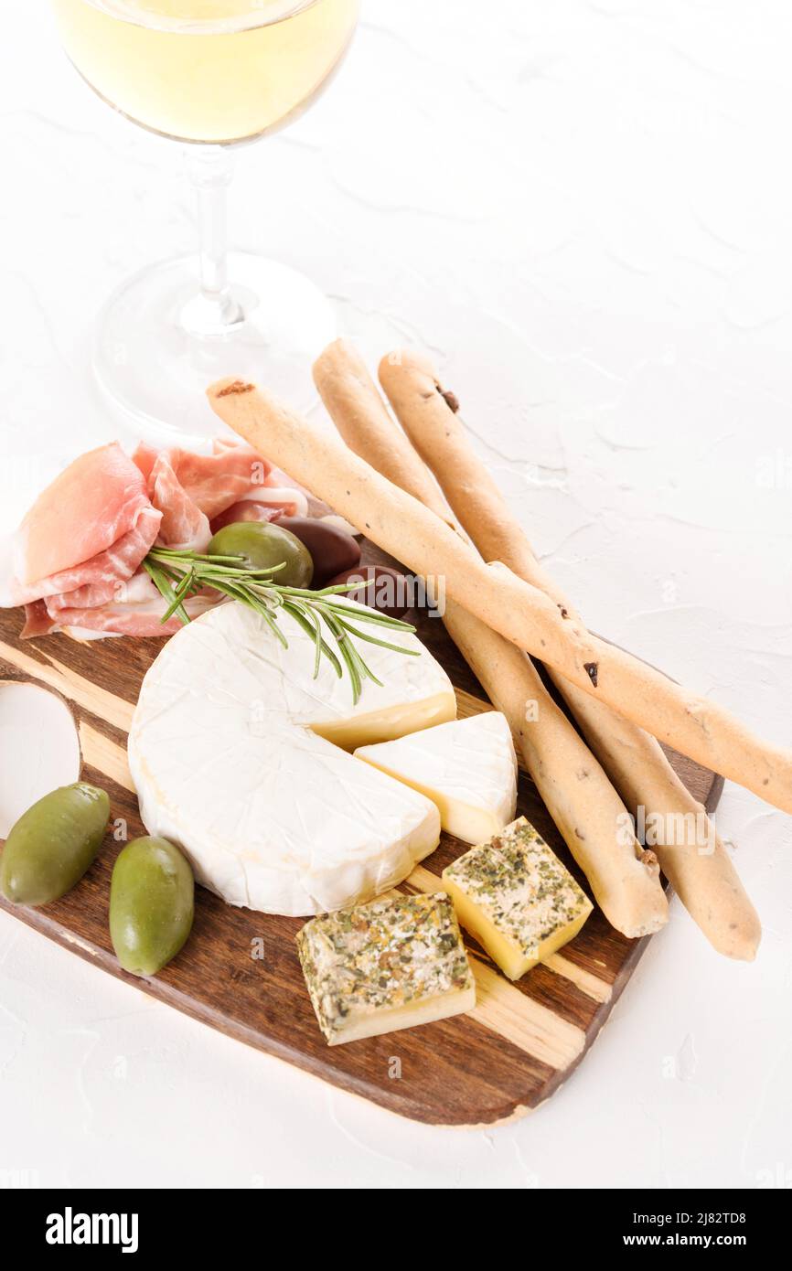 Holzbrettaufschnitt mit einer Auswahl an Käse und Prosciutto, serviert mit Oliven, Grissini und einem Glas Wein auf einem Tisch aus weißem Stein. Italienische Traditionti Stockfoto