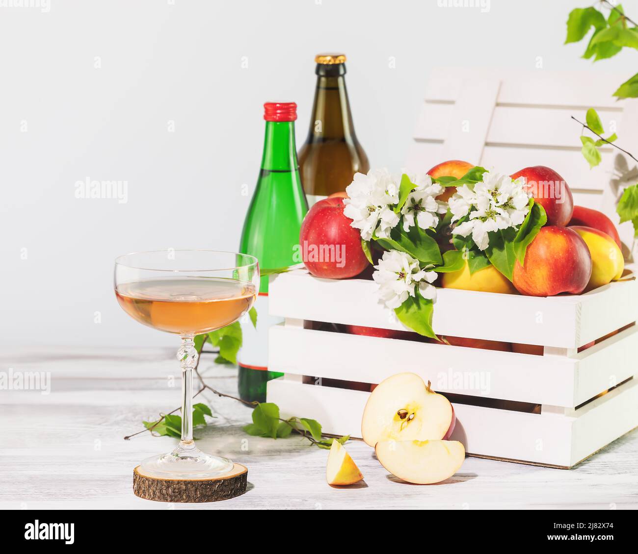 Ein Glas natürlichen Apfelmost mit Flaschen und hofeigenen Äpfeln in einer weißen Holzkiste auf neutralem Hintergrund mit Kopierraum. Sommerlich erfrischend Low-Alcoh Stockfoto