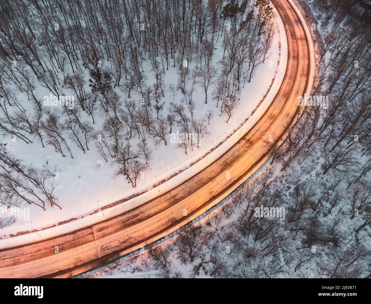 Fliegen Sie über die winterlich verschneite, leere Straßenkurve. Beleuchtete abendliche Autobahn, umgeben von schneebedecktem Wald. Luftaufnahme nach unten Stockfoto