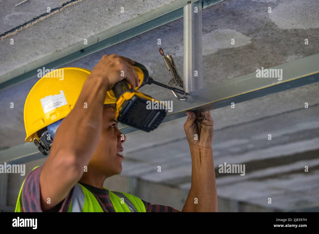 Facharbeiter im Bauwesen, der eine C-Klemme und einen elektrischen Schraubendreher verwendet, um eine Stahlstrebe an einer Stützstrebe für eine abgehängte Decke in einem zu befestigen Stockfoto