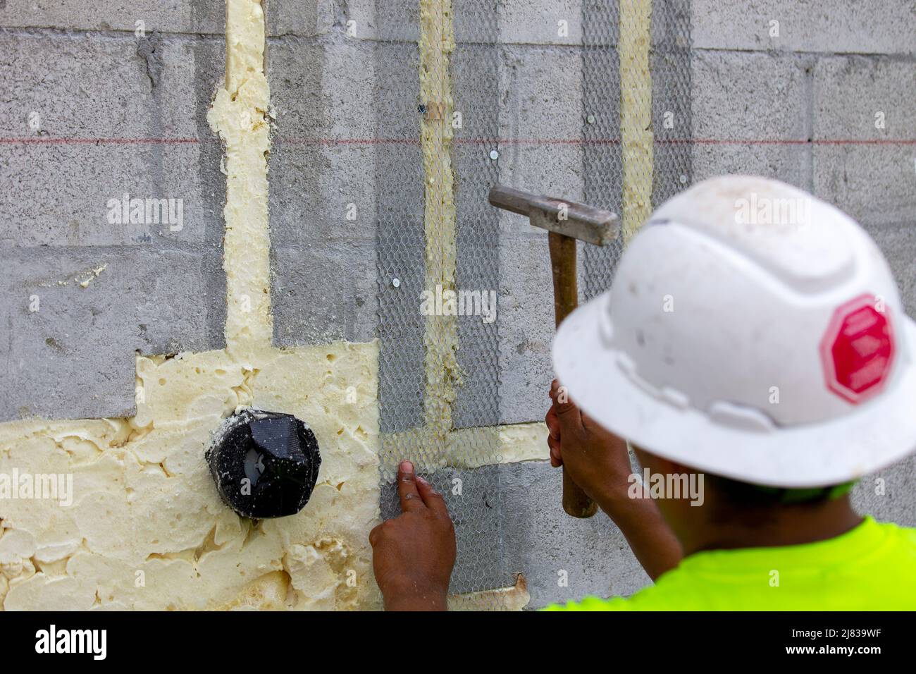 Ein Bauarbeiter befestigt ein Stahlgeflecht für den Stuckauftrag mit einem Hammer und Betonzapfen, um Rohrleitungen zu verbergen Stockfoto