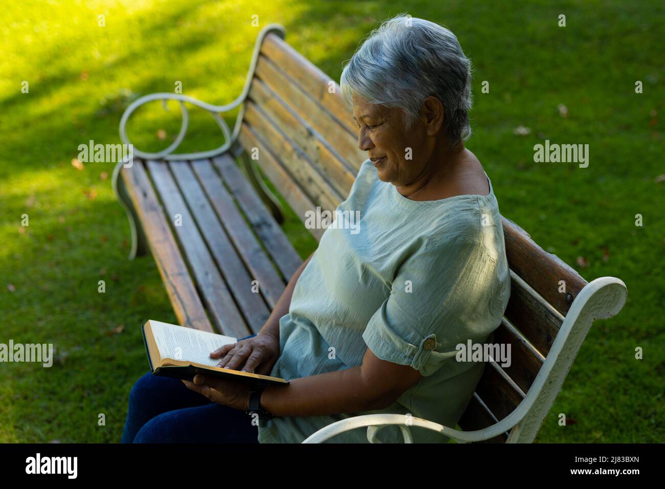 Seitenansicht einer lächelnden älteren Frau aus der Birazialzeit mit kurzen Haaren und Lesebuch auf der Bank im Park Stockfoto