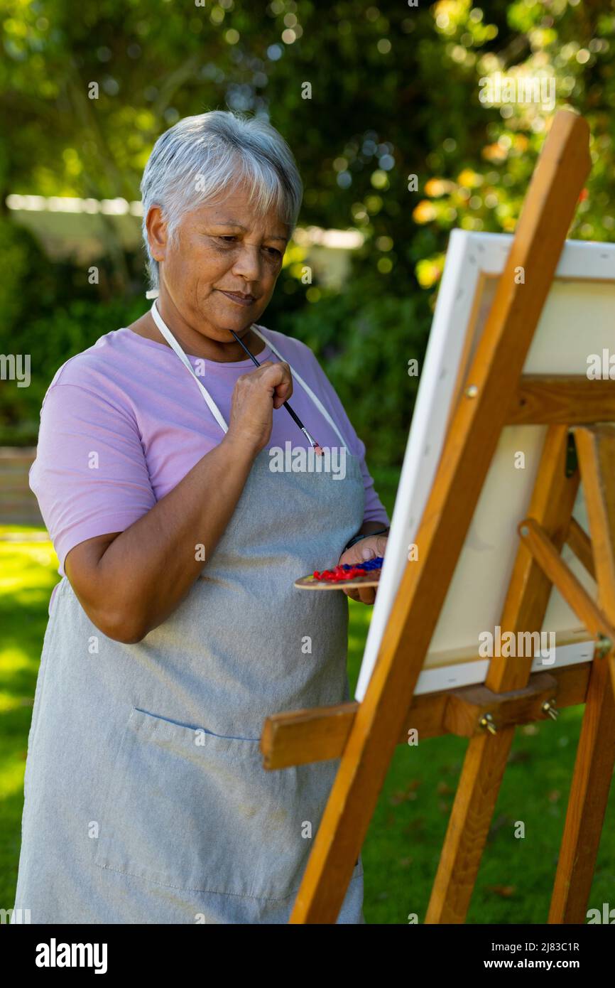 Biracial ältere Frau mit kurzen Haaren denken, während Blick auf Malerei auf Leinwand im Hof Stockfoto