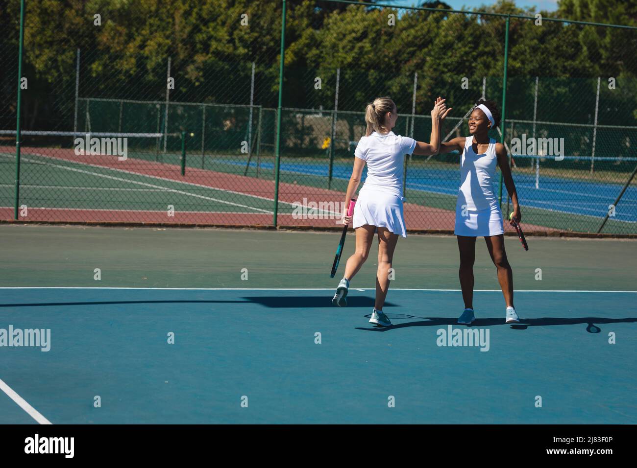 Glückliche multirassische weibliche Doppel-Team geben High-Five beim Tennisspielen auf dem Platz an sonnigen Tag Stockfoto