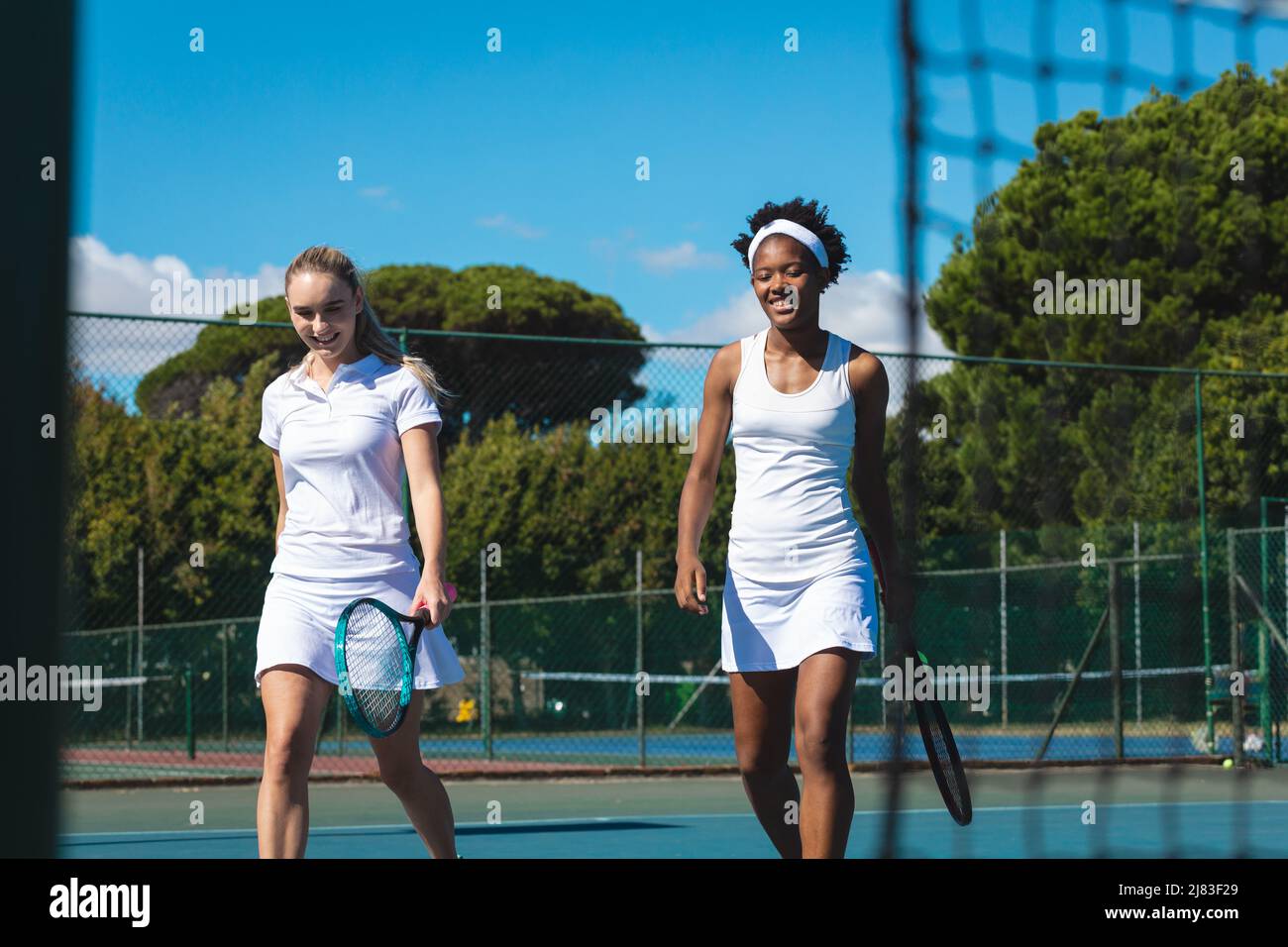 Lächelnde junge Frauen im Biracial Tennis verdoppelt das Team, das am sonnigen Tag gemeinsam auf dem Platz läuft Stockfoto