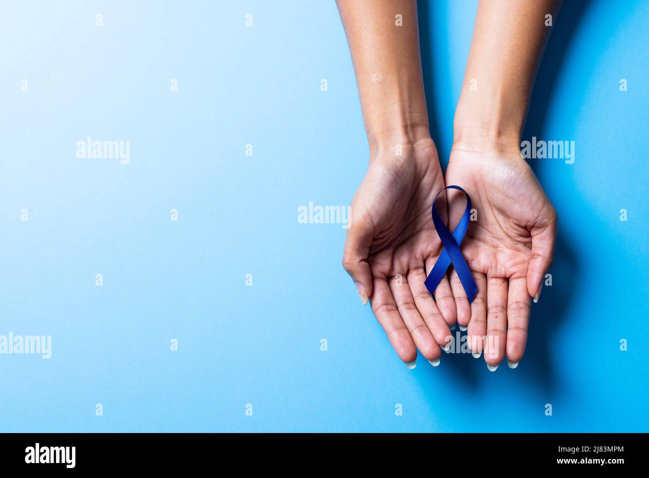 Beschnittene Hände einer afroamerikanischen Frau mit mittlerem Erwachsenen, die ein blaues Bewusstseinsband auf blauem Hintergrund hält Stockfoto