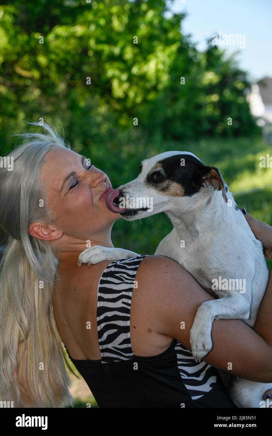 Im Garten steht jungen blonde junge junge Frau wird vom Hund im Arm mit ausgesprochter Zunge gekuesst Stockfoto