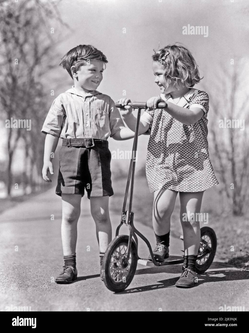 1930S 2 KINDER REDEN AUF BÜRGERSTEIG MÄDCHEN STEHEN MIT DEN HÄNDEN AUF LENKER VON SCOOTER JUNGEN IN SHORTS LÄCHELND HALTEN GIRL’S ARM - J8285 HAR001 HARS KOPIEREN RAUM FREUNDSCHAFT GANZKÖRPERPERSONEN MÄNNER TRANSPORT B&W SOMMERZEIT GLÜCK FRÖHLICH ABENTEUER BEZIEHUNGEN LÄCHELT VERBINDUNG FRÖHLICHE STILVOLLE ROLLER WACHSTUM LENKER SAISON ZWEISAMKEIT SCHWARZ UND WEISS KAUKASISCHEN ETHNIE HAR001 ALTMODISCH Stockfoto