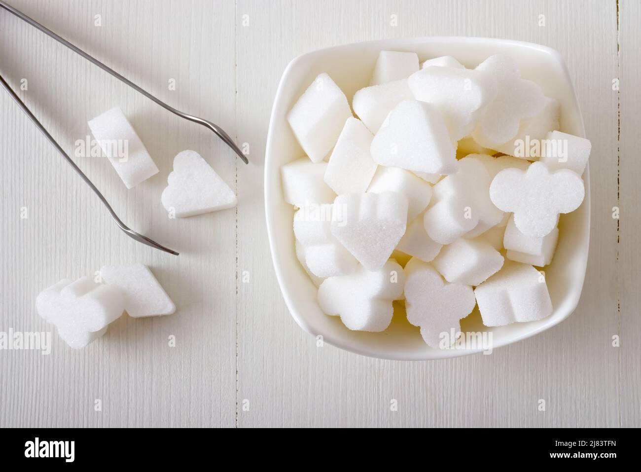 Brückenförmiger Rübenzucker in Porzellanschale auf einem weißen Holztisch mit silberner Zuckerzange. Flach liegend, Blick von oben nach unten, keine Leute. Stockfoto