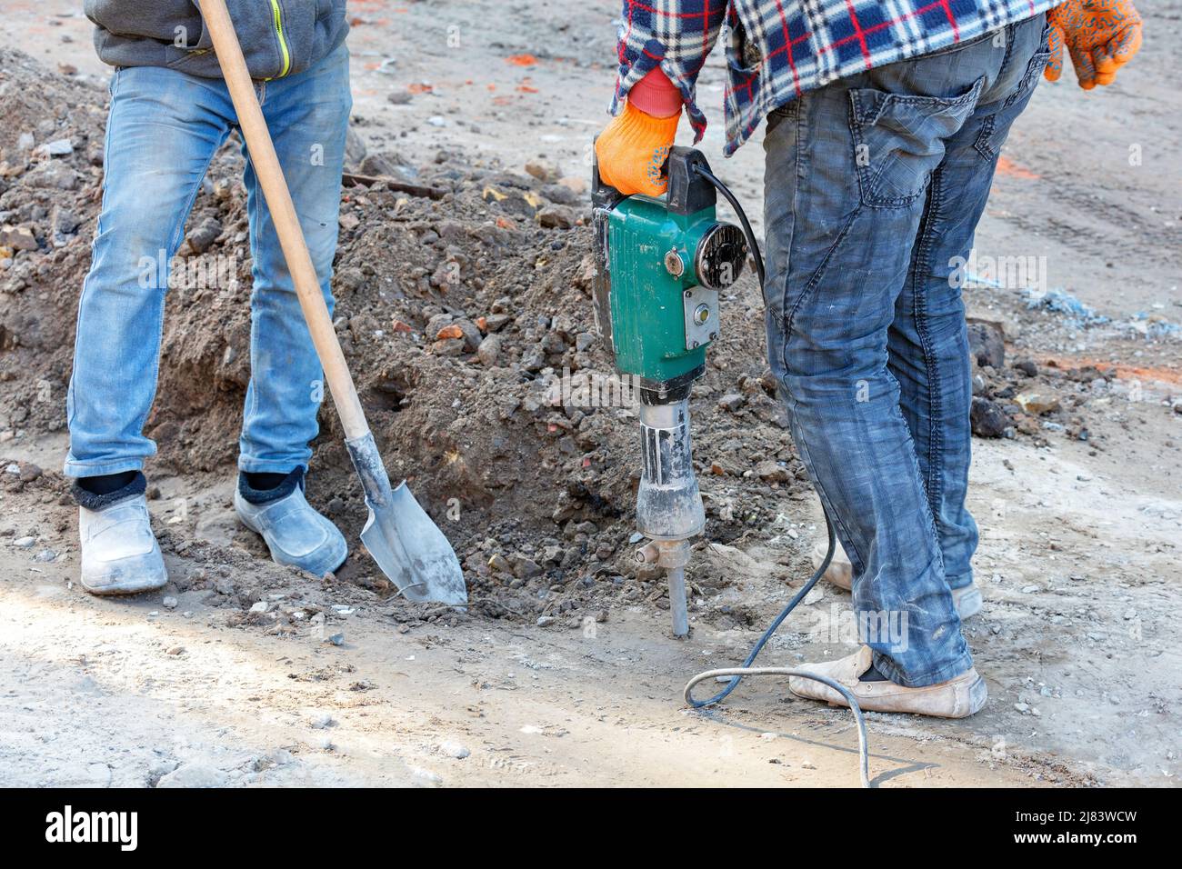 Arbeiter in blauen Jeans lockern den sandigen Boden für den Bau eines Grabens auf einer Baustelle mit einem elektrischen Presslufthammer und einer Schaufel. Stockfoto