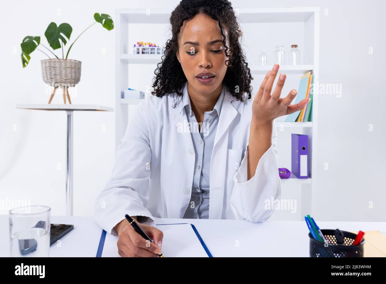 afroamerikanische Ärztin mit mittlerem Erwachsenen, die gestikuliert, während sie am Schreibtisch im Krankenhaus auf Papier schrieb Stockfoto