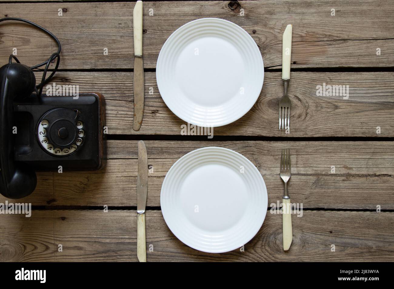 Ein altes Retro-Telefon und zwei Teller mit Messern und Gabeln auf einem Holztisch in der Küche, telefonische Bestellung von Speisen zum Abendessen, ein Retro-Telefon auf dem Tisch Stockfoto