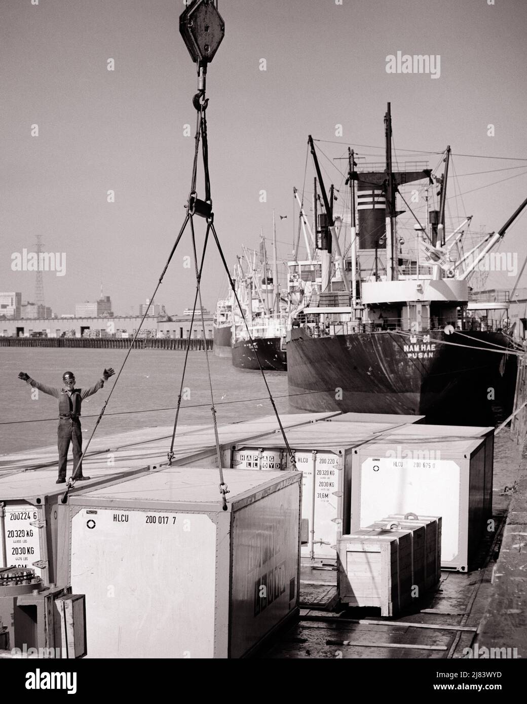 1970S ANONYME MANN-WAFFEN, DIE DAS ANHEBEN EINES FRACHTCONTAINERS AUF ODER VON EINEM CONTAINERSCHIFF LEITEN SCHIFFE AM DOCK - S17386 HAR001 HARS SCHIFFE VERTRAUEN GESTIK TRANSPORT FRACHT B&W ZIELE CONTAINER GESCHICKLICHKEIT BERUF FÄHIGKEITEN HOHEN WINKEL RICHTUNG ARBEIT BESCHÄFTIGUNG GESTEN BERUFE VERBINDUNG INFRASTRUKTUR ANONYME MITARBEITER FRACHTER ODER ZUSAMMENARBEIT REGIE PRÄZISION VERSAND LIEFERN BINNENSCHIFF SCHWARZ UND WEISS KAUKASISCHE ETHNIE HAR001 WERKTÄTIGEN ALTMODISCHEN SCHIFF Stockfoto