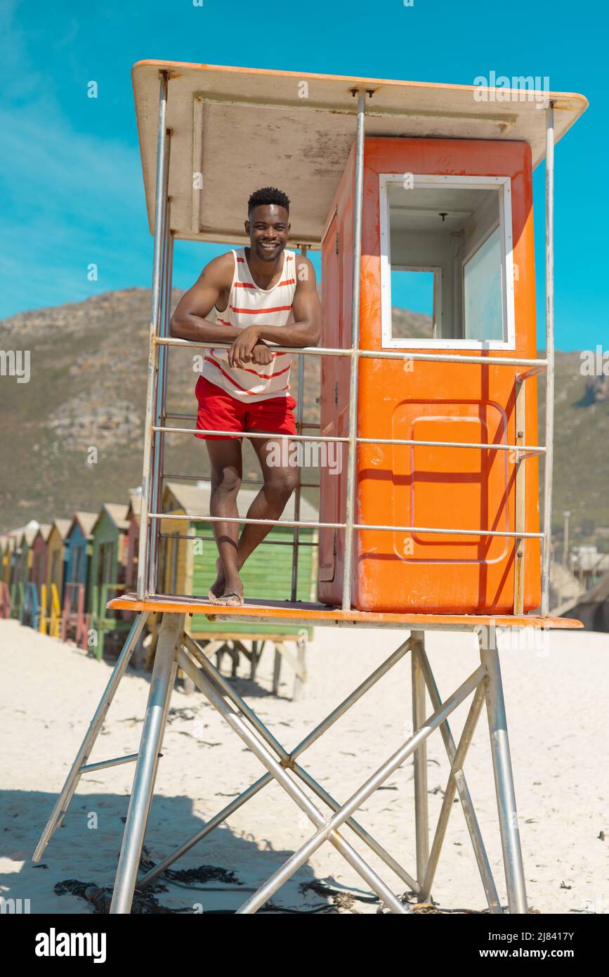 Porträt eines lächelnden afroamerikanischen jungen Mannes, der am Geländer auf der Rettungsschwimmerhütte gegen den blauen Himmel steht Stockfoto