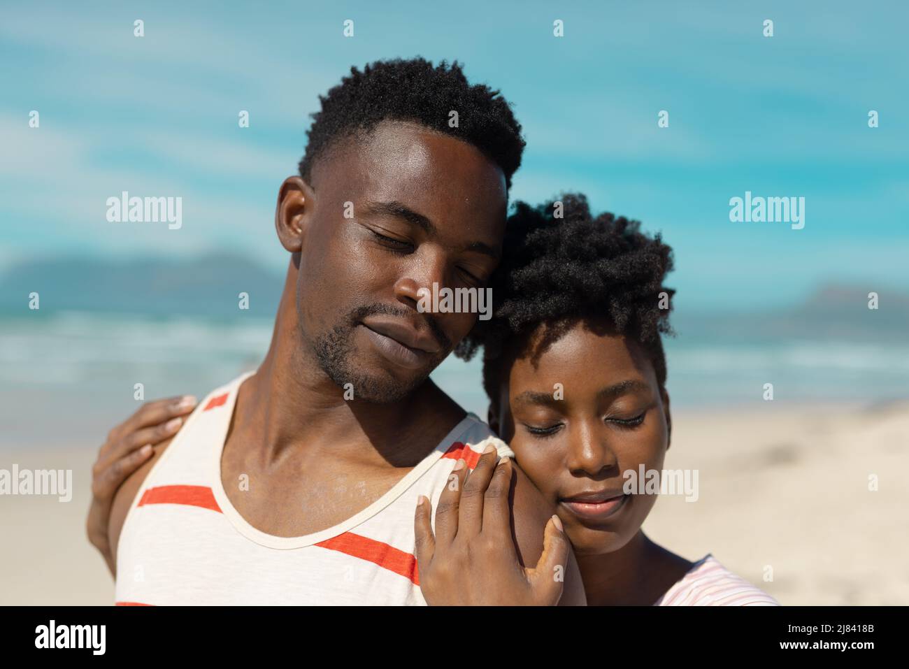 Liebende afroamerikanische junge Frau mit geschlossenen Augen umarmt Freund am Strand gegen blauen Himmel Stockfoto