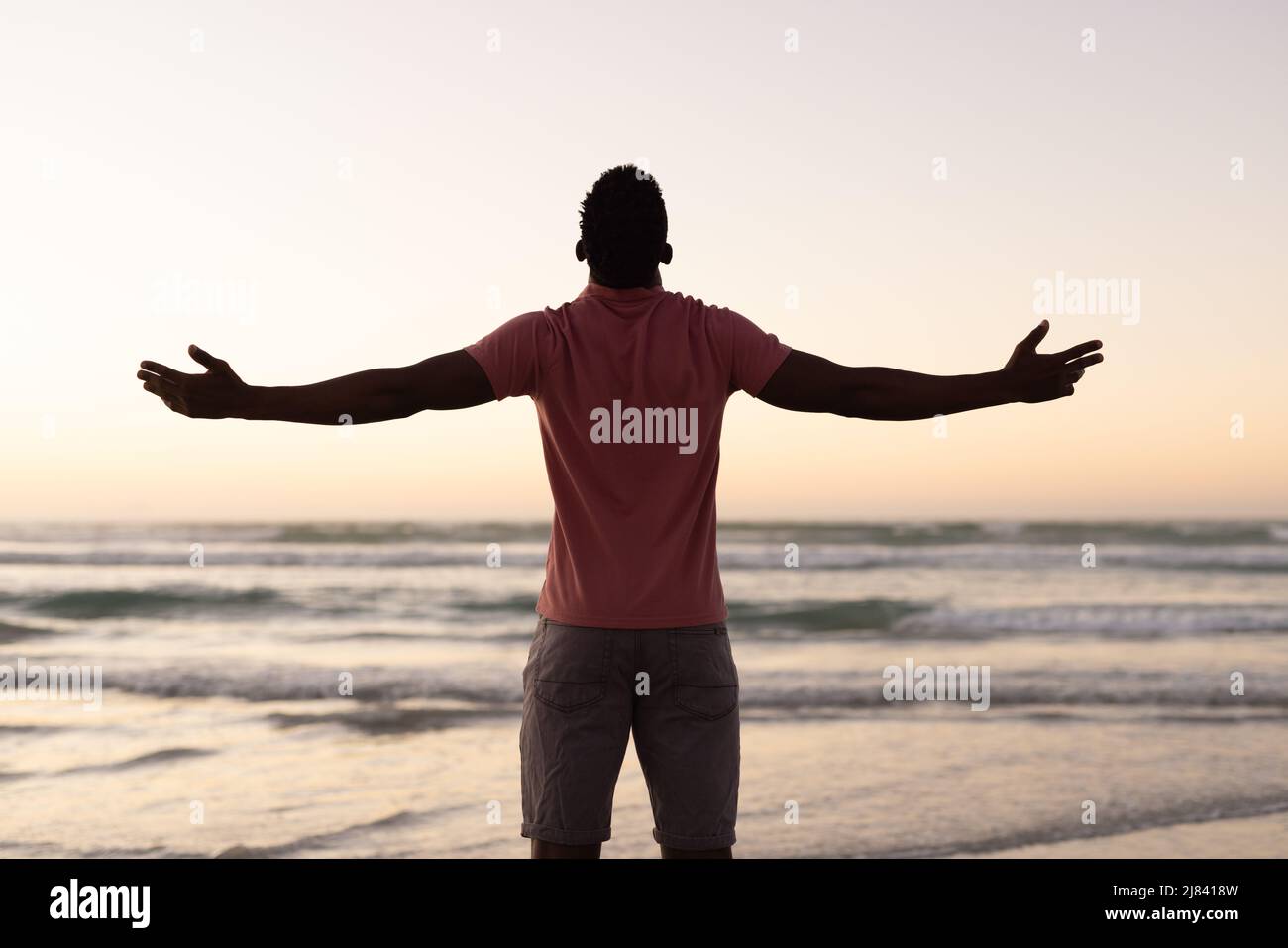 Rückansicht eines sorglosen afroamerikanischen jungen Mannes mit ausgestreckten Armen am Strand vor klarem Himmel Stockfoto