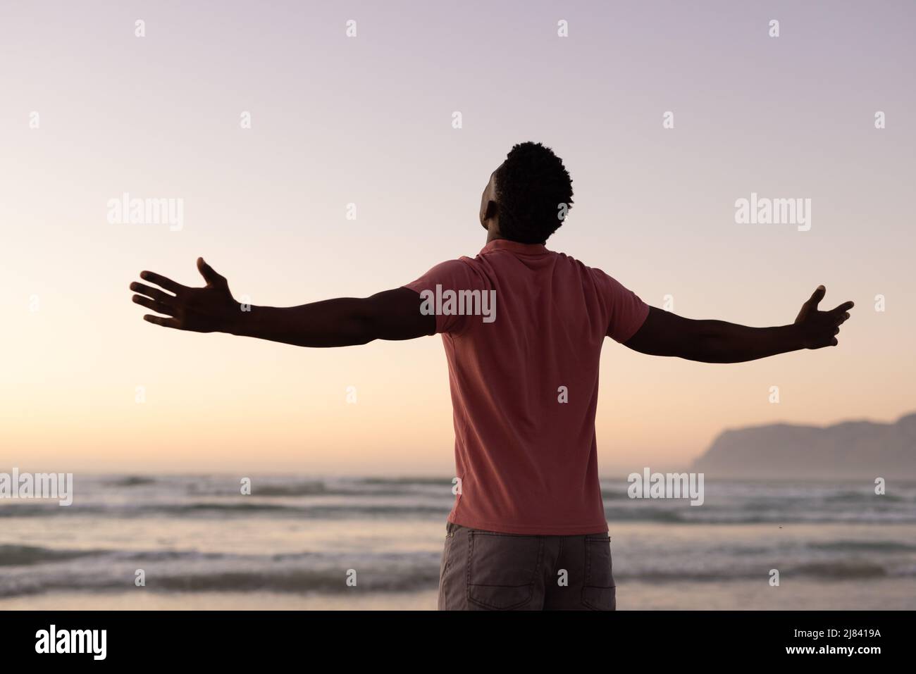 Rückansicht eines sorglosen afroamerikanischen jungen Mannes mit ausgestreckten Armen, der bei Sonnenuntergang am Strand stand Stockfoto