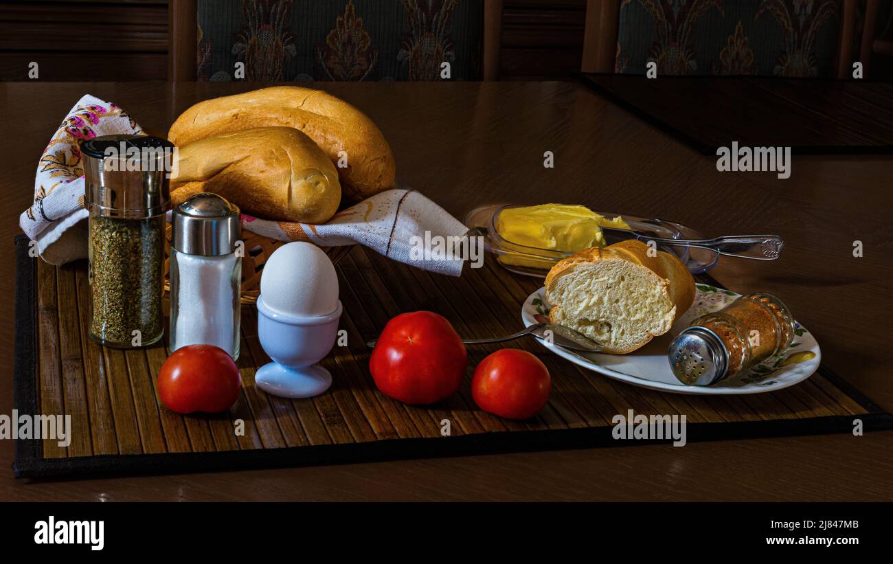 Ein weich gekochtes Ei zum Frühstück mit frischen Brötchen, Butter, einer Tasse Tee und einem Haufen schöner Tomaten. Das Foto kann auf Leinwand und Poster verwendet werden. Stockfoto