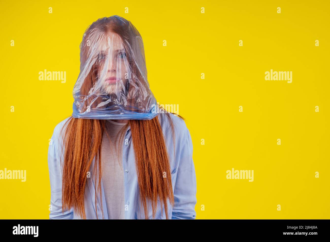 Rothaarige Ingwer europäische Frau Erstickungskopf in blauen Polyethylen-Paket in Studio gelben Hintergrund. Eco Verschmutzung Cellophan Film Konzept Stockfoto