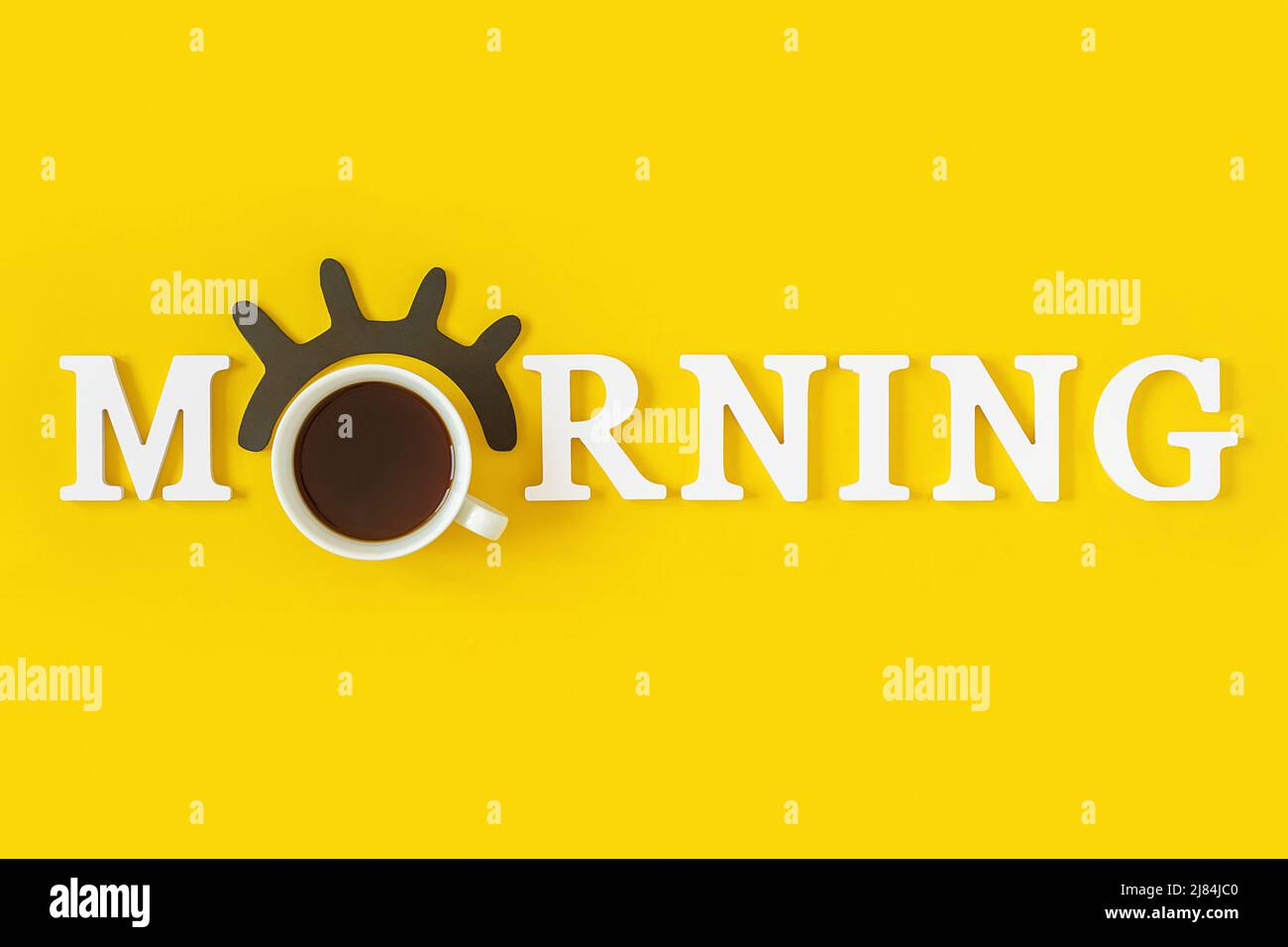 Kreatives Good Morning-Konzept. Weiße Buchstaben, schwarze Wimpern, Wecker und eine Tasse Kaffee auf gelbem Hintergrund. Draufsicht, Flat Lay. Grußkarte. Stockfoto