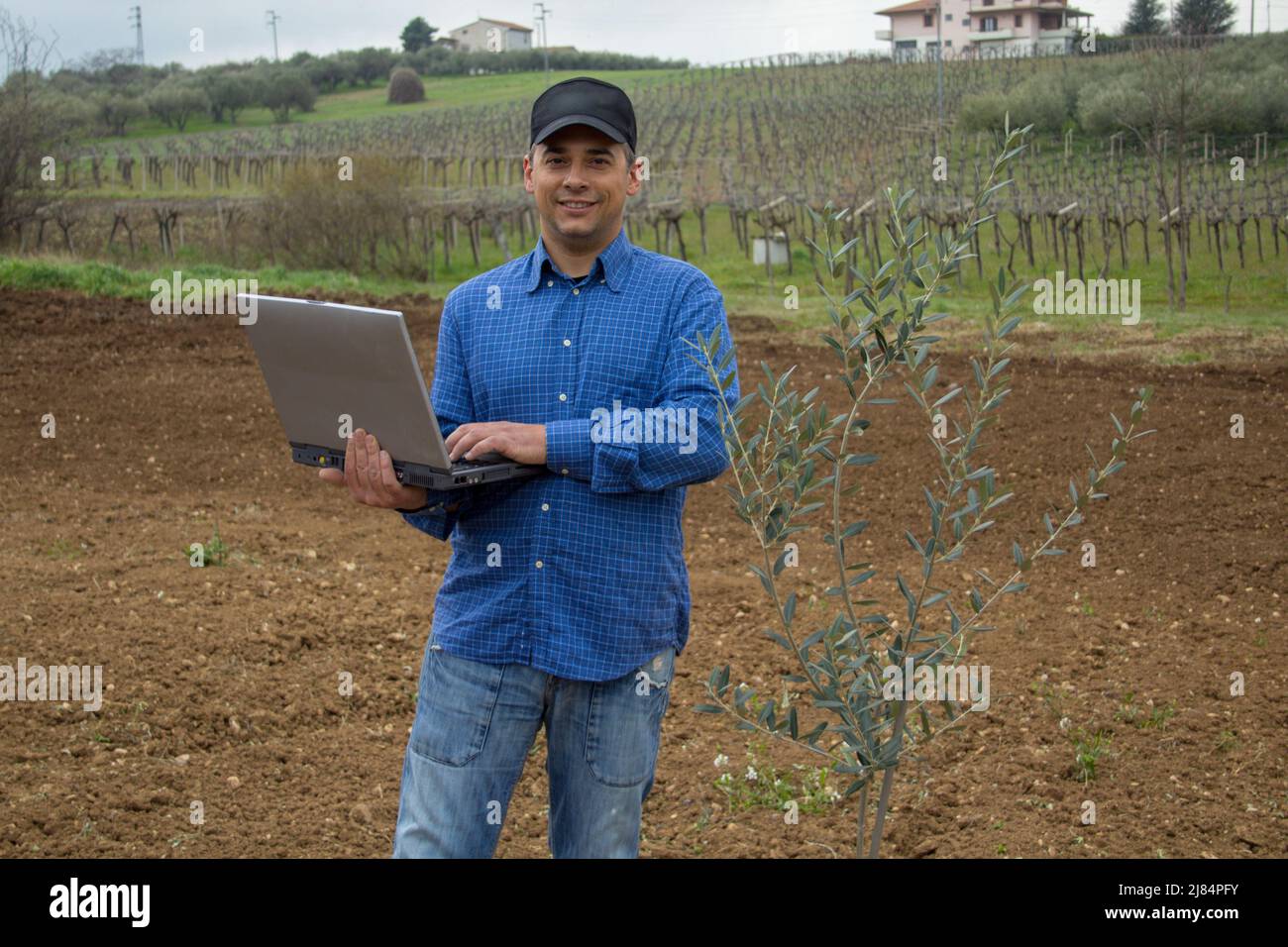 Foto eines lächelnden Bauern mit einem Laptop in der Hand, nachdem er einen Olivenbaum auf einem Feld gepflanzt hat. Stockfoto