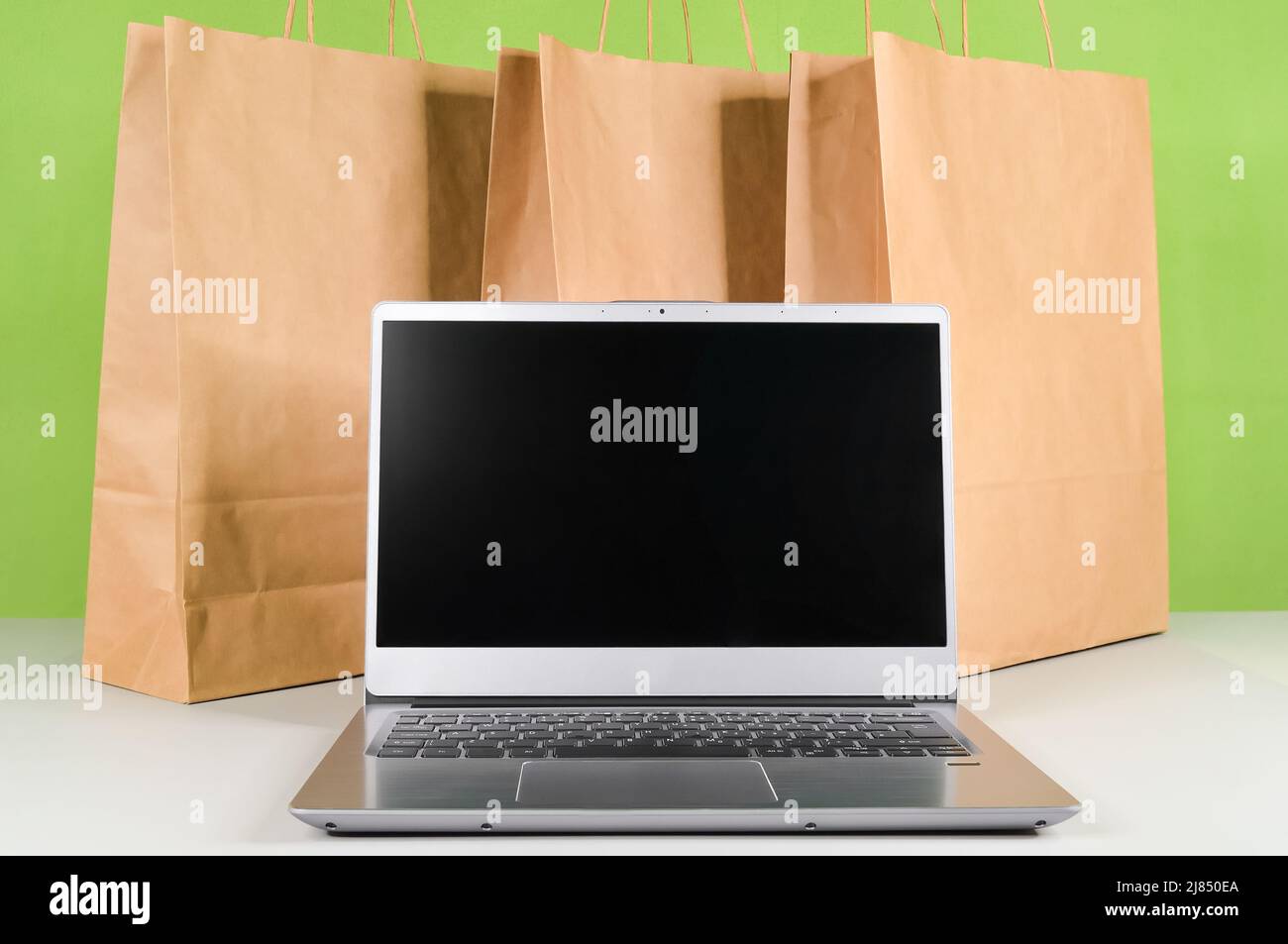 Laptop-Computer mit leerem Bildschirm vor dem Hintergrund von Papiertüten mit Waren und Einkäufen, die auf dem Online-Markt bestellt und nach Hause geliefert wurden Stockfoto