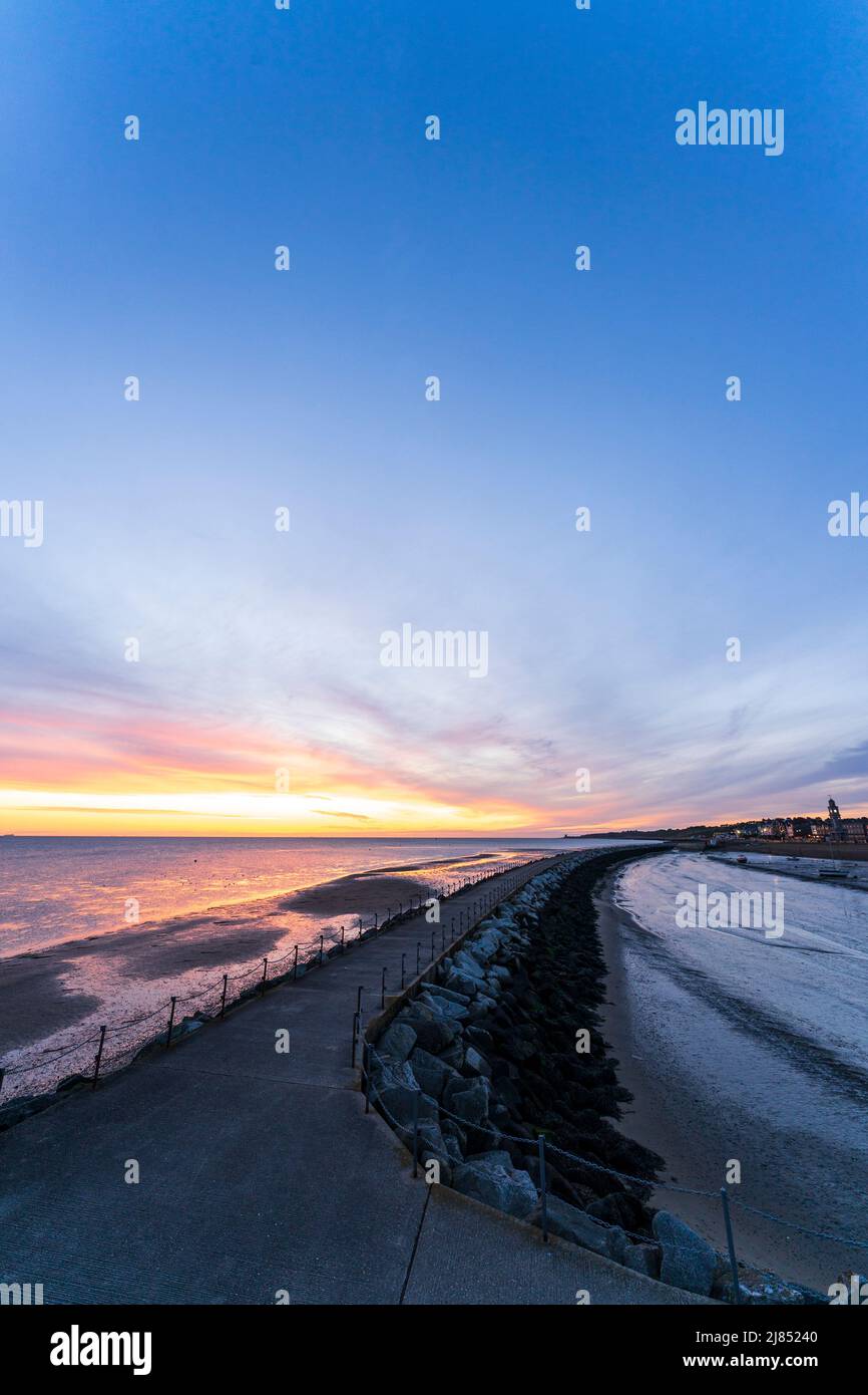 Der Morgenhimmel über der Nordsee, bei Ebbe vom Hafen Herne Bay in Kent aus gesehen. Die Hafenmauer in der Mitte erstreckt sich auf der einen Seite bis zur Stadtpromenade und auf der anderen Seite bis zum orangen Himmel über dem Meer. Wolkiger Himmel, aber klares oranges Band am Horizont. Stockfoto