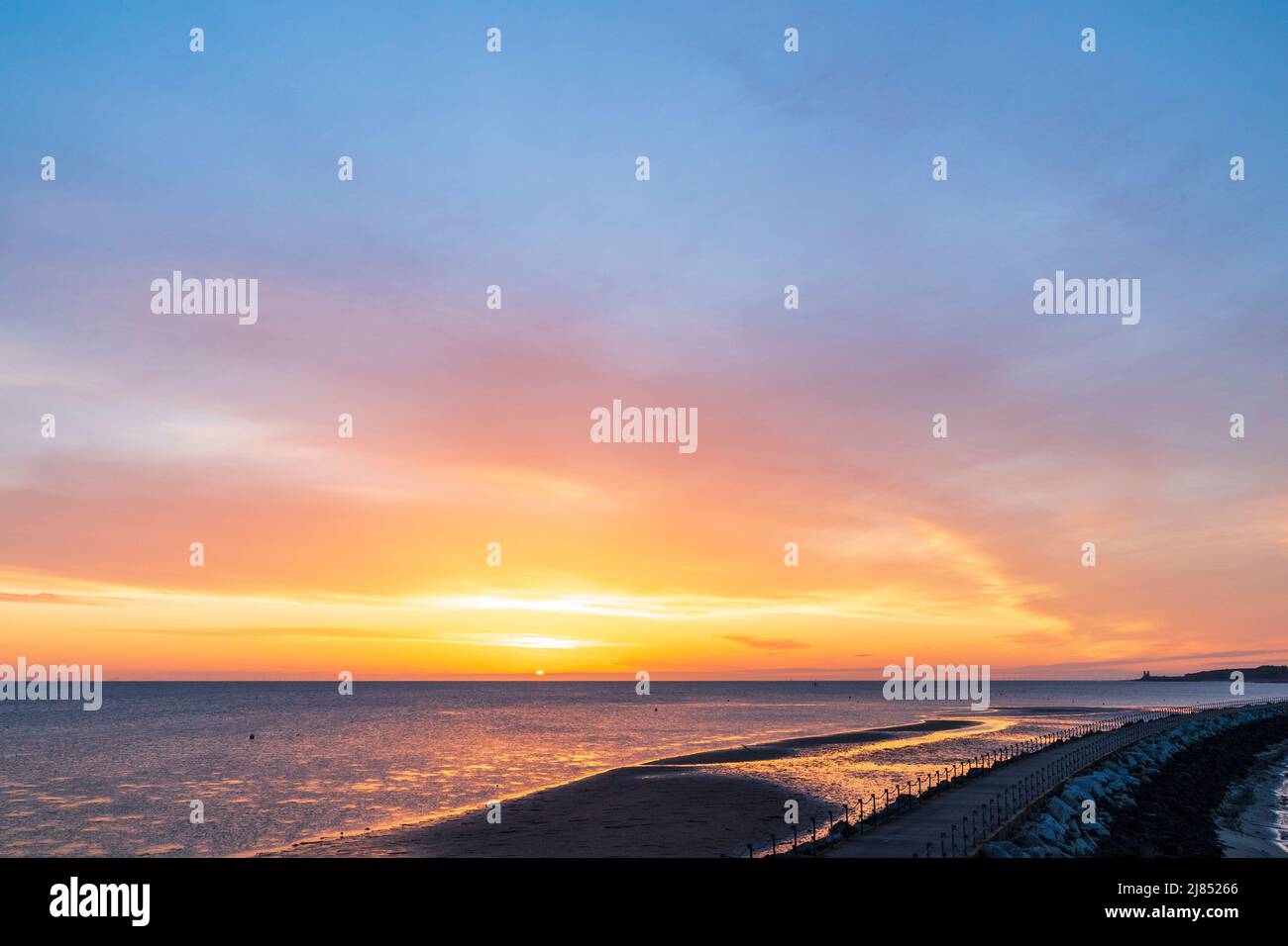 Der Morgenhimmel über der Nordsee, bei Ebbe vom Hafen Herne Bay in Kent aus gesehen. Die Hafenmauer in der Mitte erstreckt sich auf der einen Seite bis zur Stadtpromenade und auf der anderen Seite bis zum orangefarbenen Morgenhimmel über dem Meer. Wolkiger Himmel, aber klares oranges Band am Horizont. Stockfoto