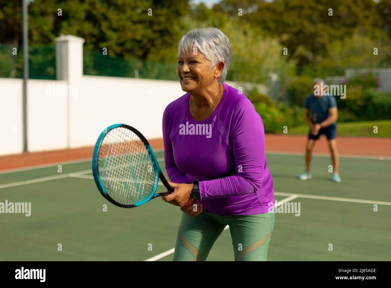 Lächelnde ältere Frau aus der Birazialzeit mit kurzen Haaren, die auf dem Tennisplatz mit einem älteren Mann im Hintergrund Tennis spielt Stockfoto
