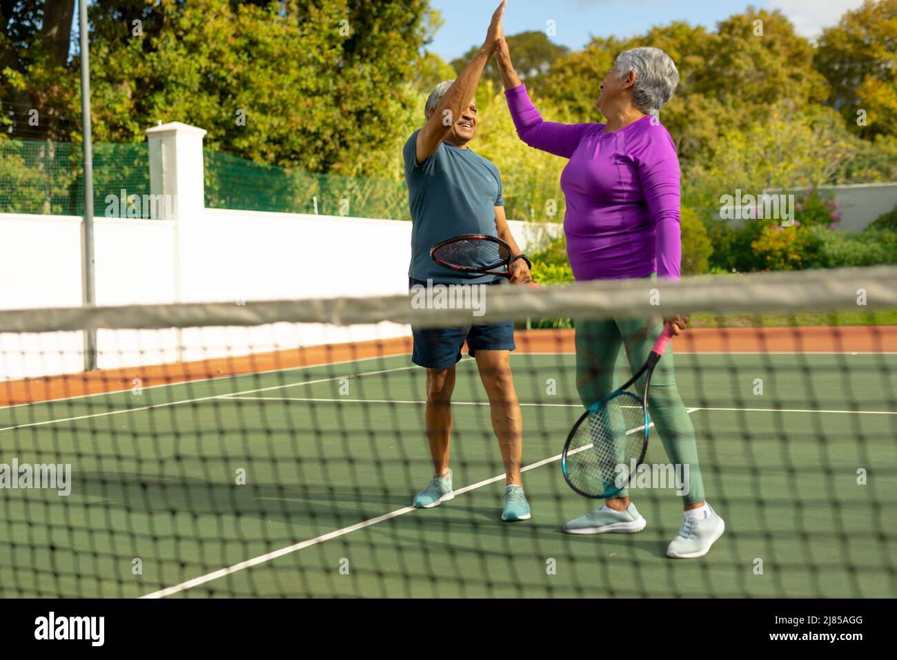 Fröhliches Senioren-Paar mit Biracial, das Schläger hält und auf dem Tennisplatz gegen Bäume High-Five gibt Stockfoto