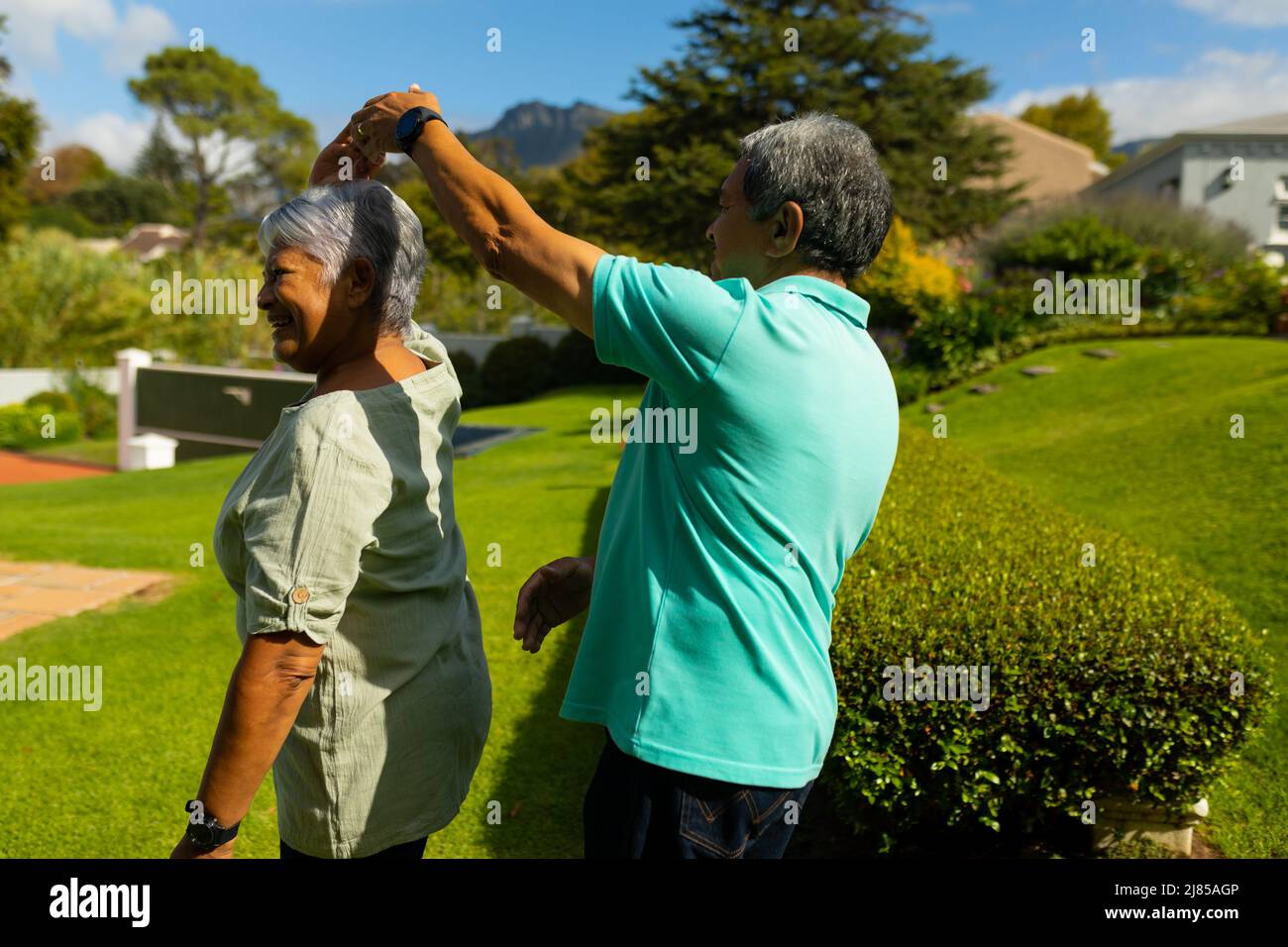 Glücklicher älterer Biracial-Mann, der an sonnigen Tagen mit seiner älteren Frau auf einem grasbewachsenen Hügel im Park tanzt Stockfoto