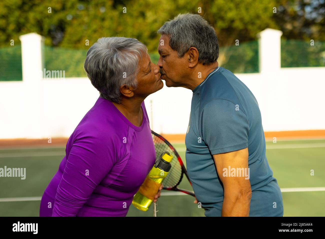 Seitenansicht eines liebenden Senioren-Paares mit geschlossenen Augen, die sich küssen, während sie auf dem Tennisplatz stehen Stockfoto