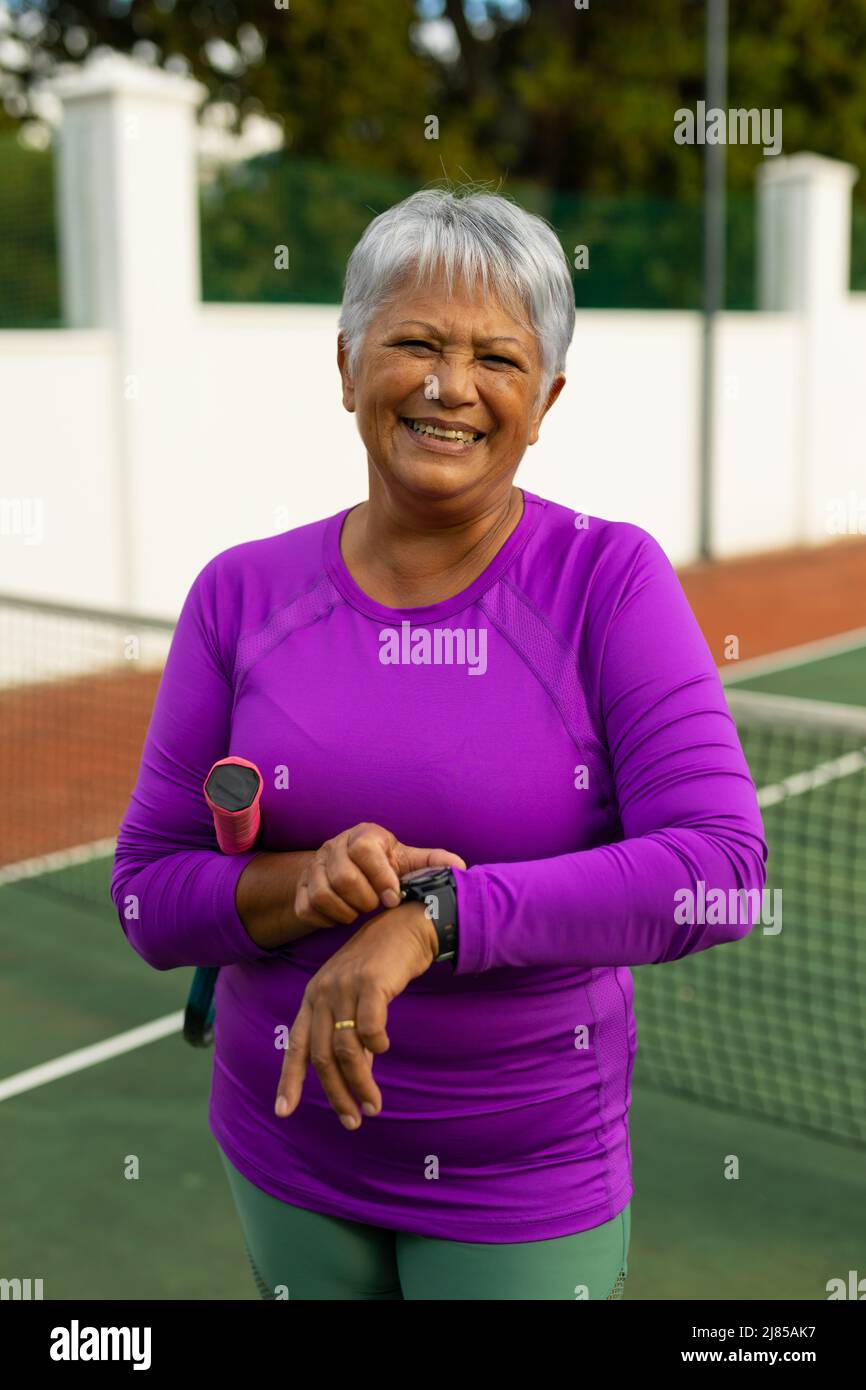 Porträt einer fröhlichen älteren Frau mit kurzer Haarprüfung über die Armbanduhr auf dem Tennisplatz Stockfoto