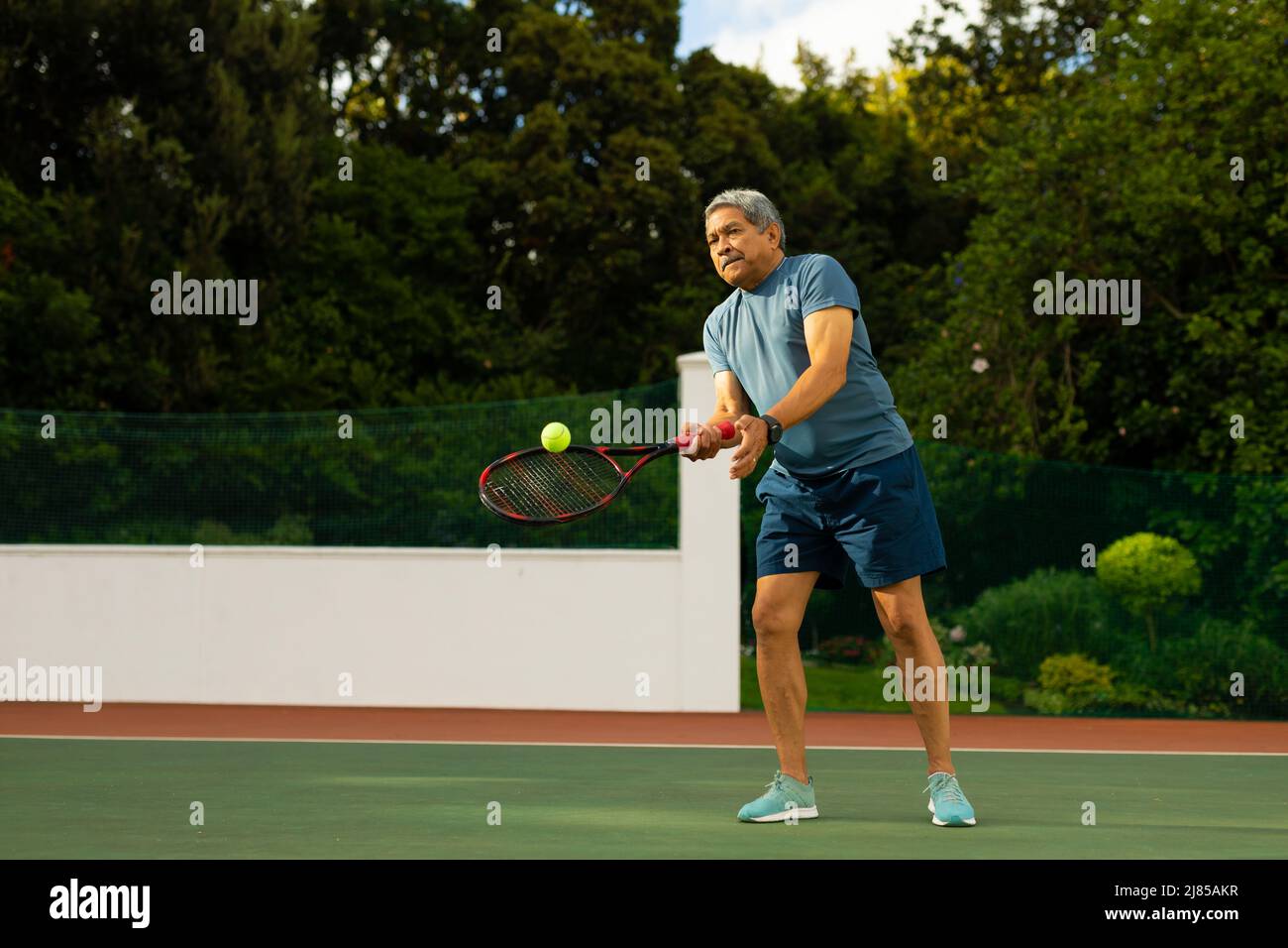 Biracial Senior Mann schlagen Tennisball mit Schläger, während Tennis gegen Bäume auf dem Platz spielen Stockfoto