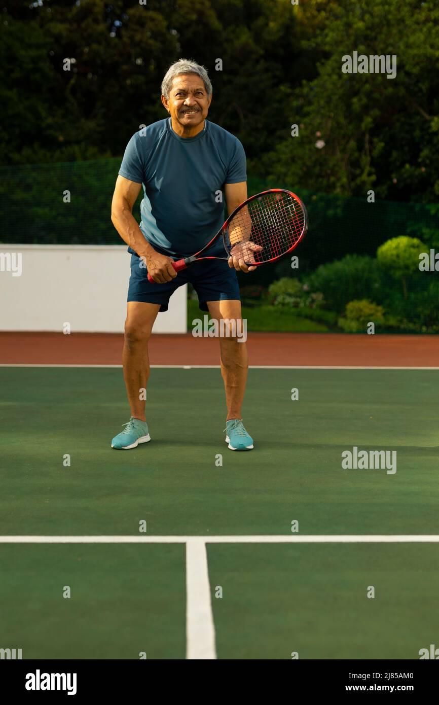 Selbstbewusster älterer Biracial-Mann mit Schläger beim Tennisspielen, während er auf dem Platz gegen Bäume steht Stockfoto