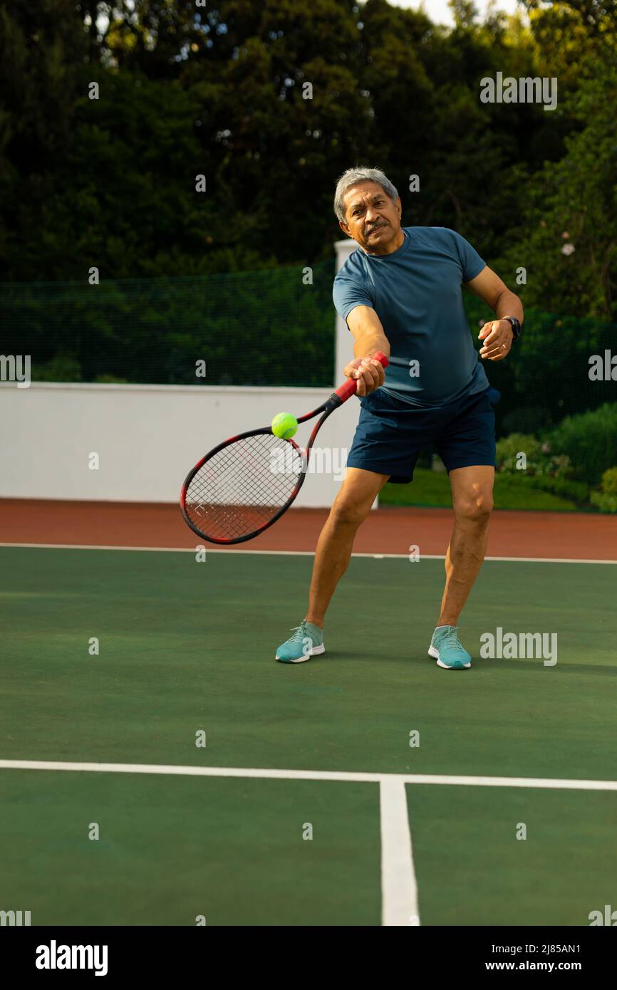 Biracial Senior Mann schlagen Tennisball mit Schläger, während Tennis gegen Bäume auf dem Platz spielen Stockfoto