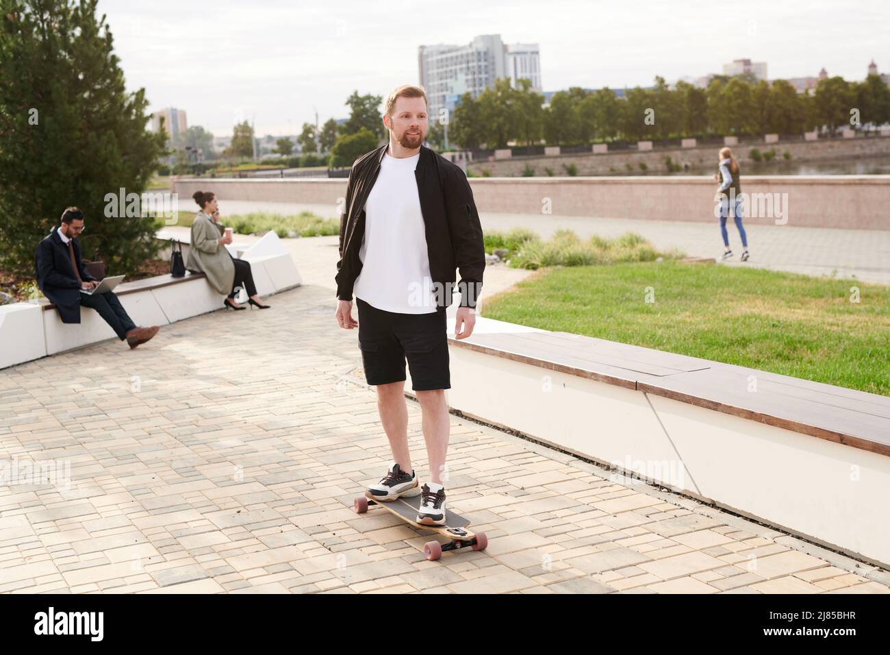 Hübscher junger bärtiger Mann in Jacke genießt Skateboarding im Stadtpark Stockfoto