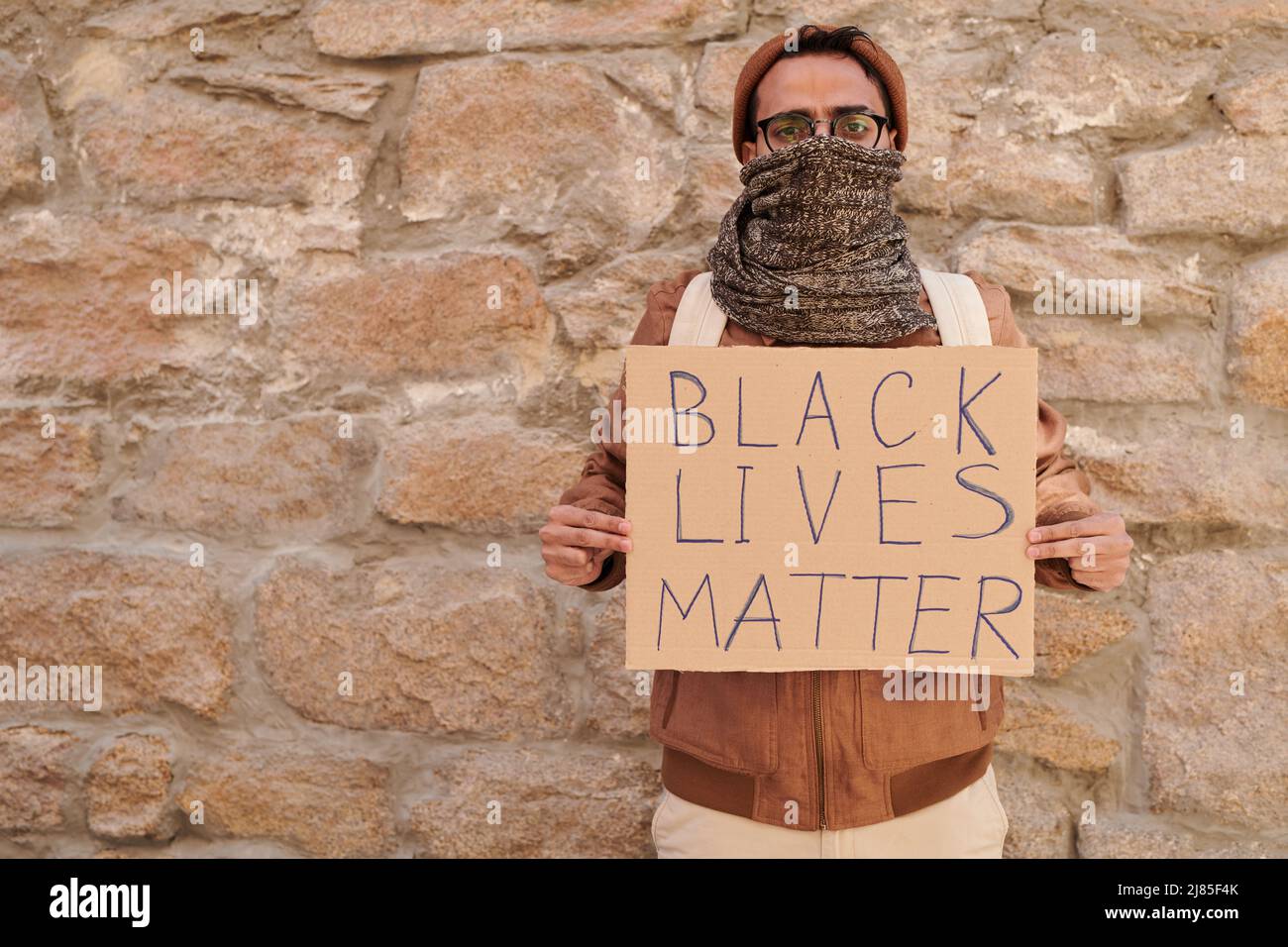 Porträt eines jungen schwarzen Mannes mit Hut und Schal, der das Banner „Black Lives Matter“ an einer alten Steinwand hält Stockfoto