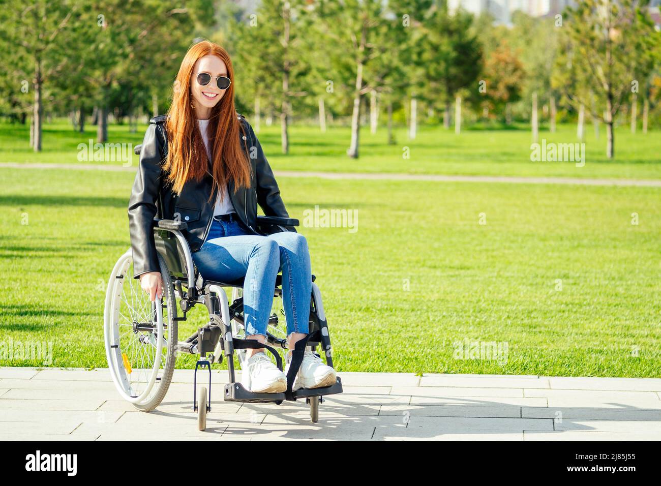 Rothaarige Ingwerfrau, die glücklich ist, sie sitzt im Rollstuhl, trägt eine Sonnenbrille und eine warme Lederjacke im Sommerpark Stockfoto